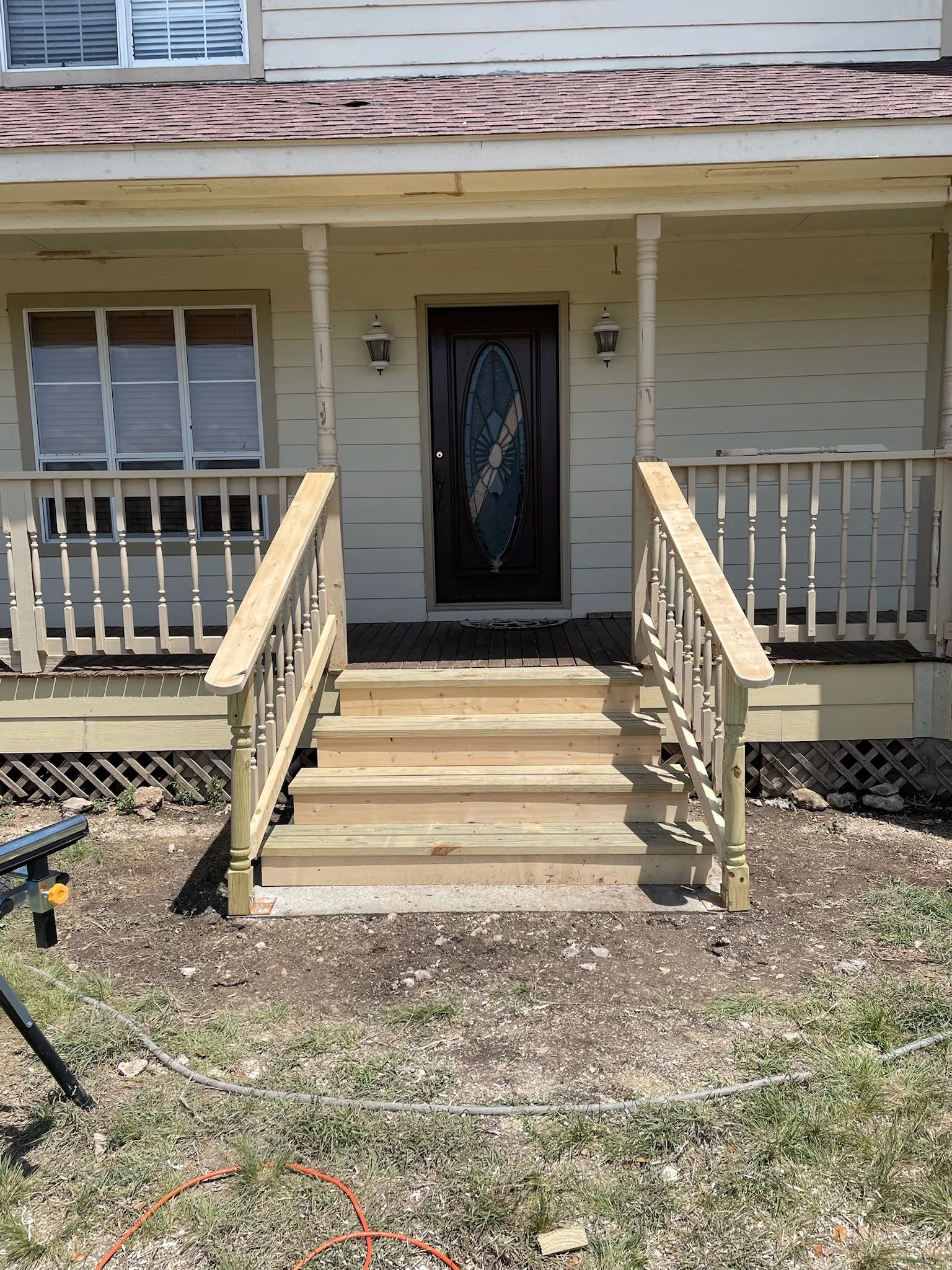A front porch with stairs and a black door on a beige house, with two lamps on either side of the door and a window to the left. The porch has new wooden railings and stairs, and the ground in front is bare soil with some grass and a garden hose.