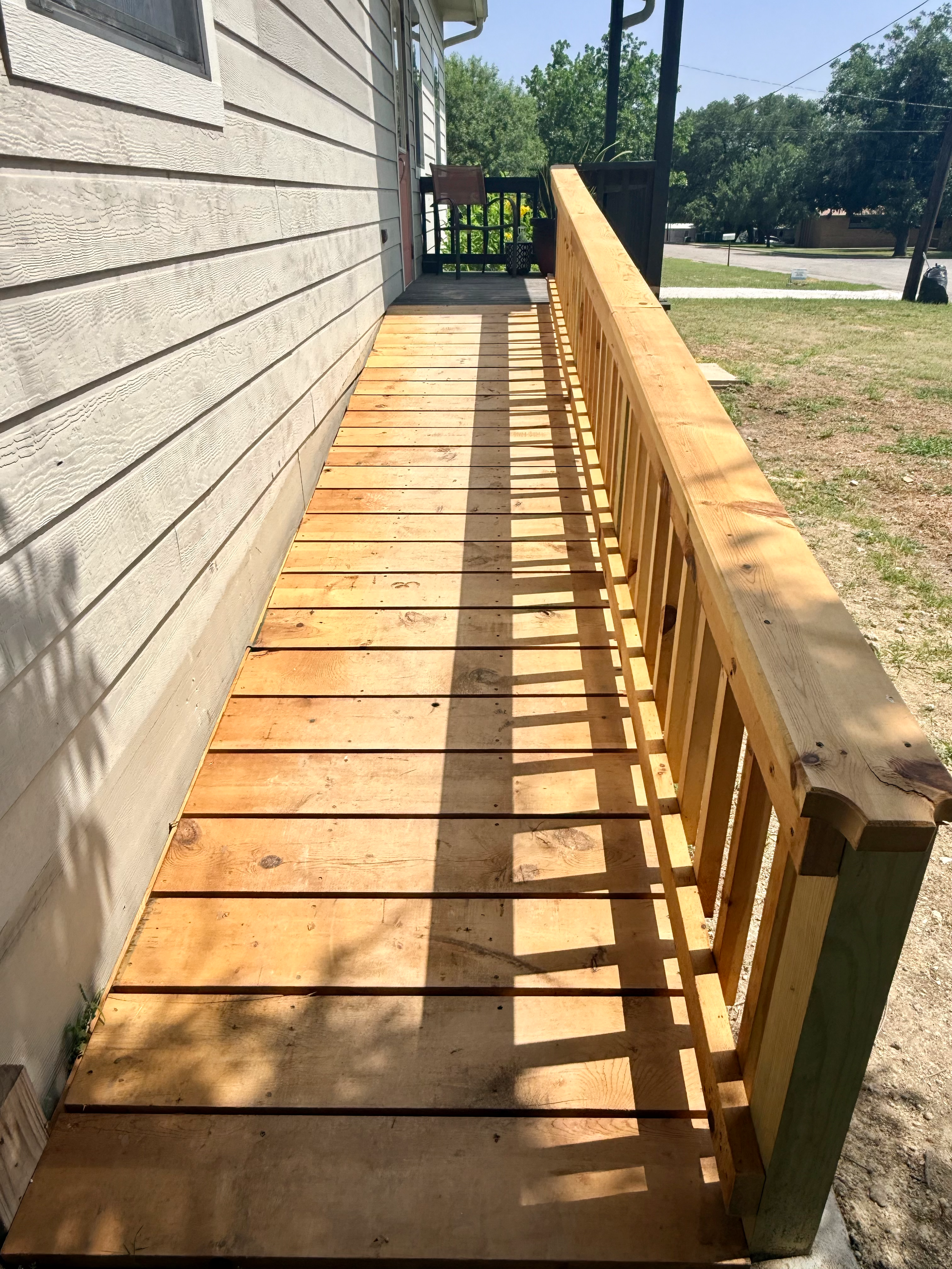 Front porch with a new wooden deck and railing adjacent to a beige house with horizontal siding, roof overhang, and backyard with trees and a street in the background.
