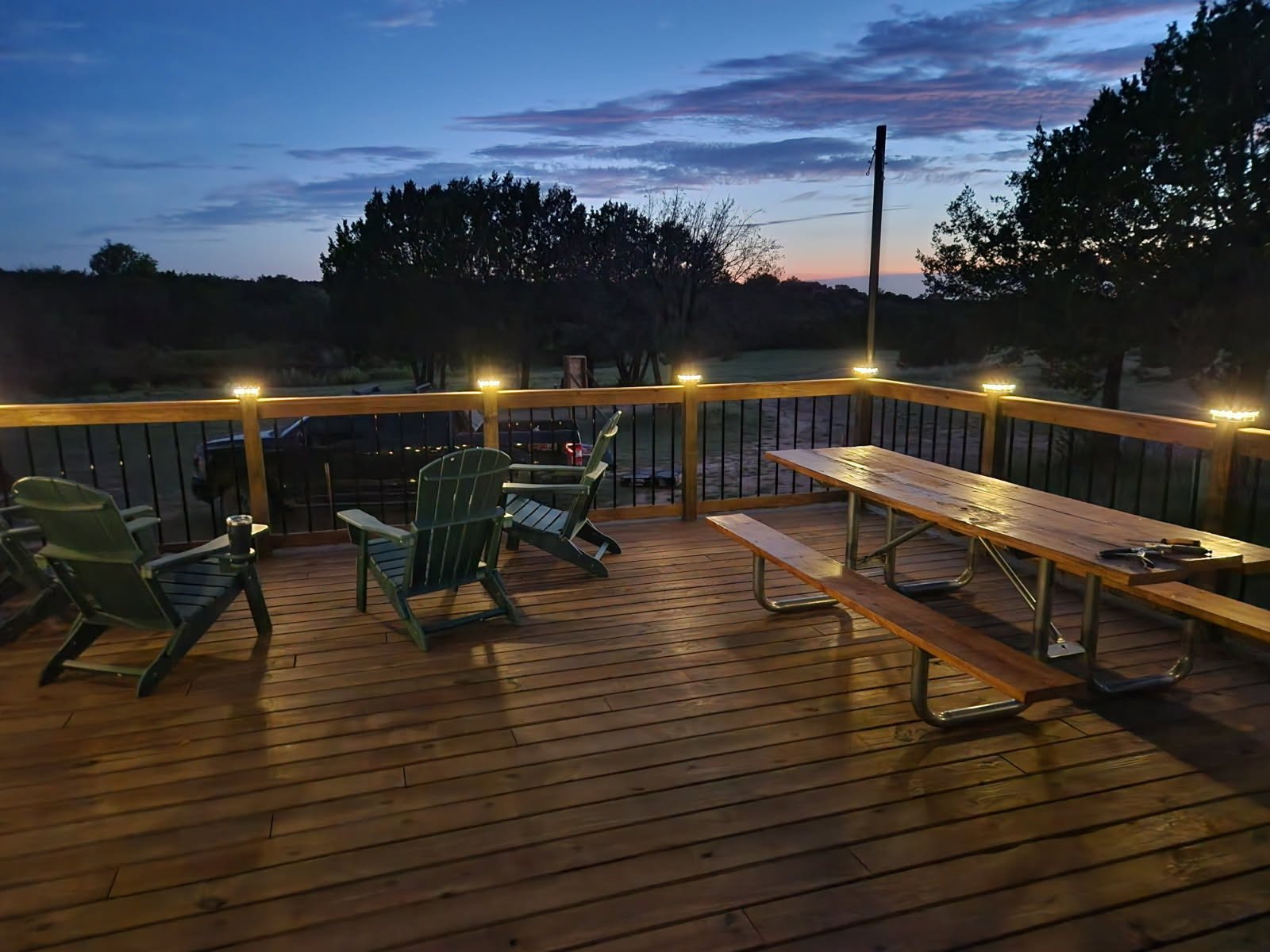 A wooden deck with Adirondack chairs, a picnic table with attached benches, and string lights along the railing. The background shows trees and a partly cloudy sky at dusk.