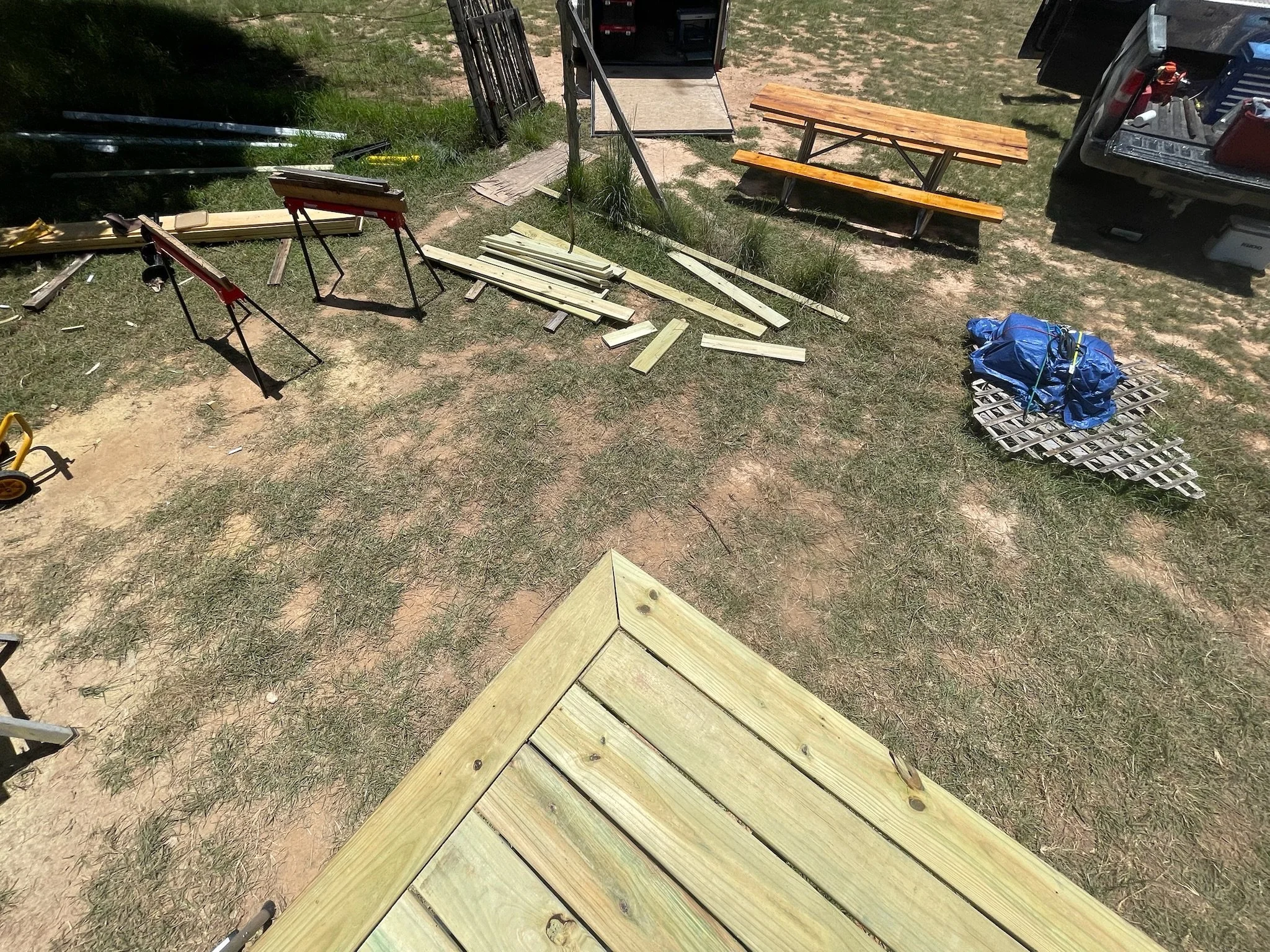 Lumber pieces, some assembled into a corner structure, woodworking tools, and a work table outdoors on a grassy area near a vehicle.