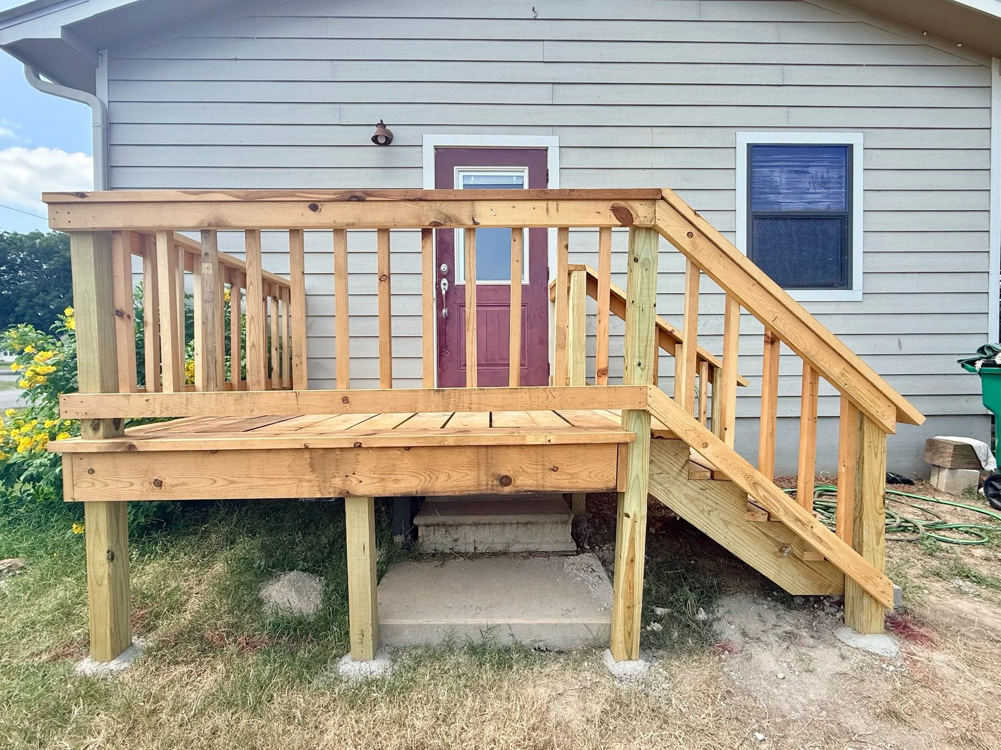 A house with gray horizontal siding and a small set of wooden stairs leading up to a purple front door. There is a window to the right of the door and a small outdoor light fixture above the door. The house has a sloped roof and the yard has some yel
