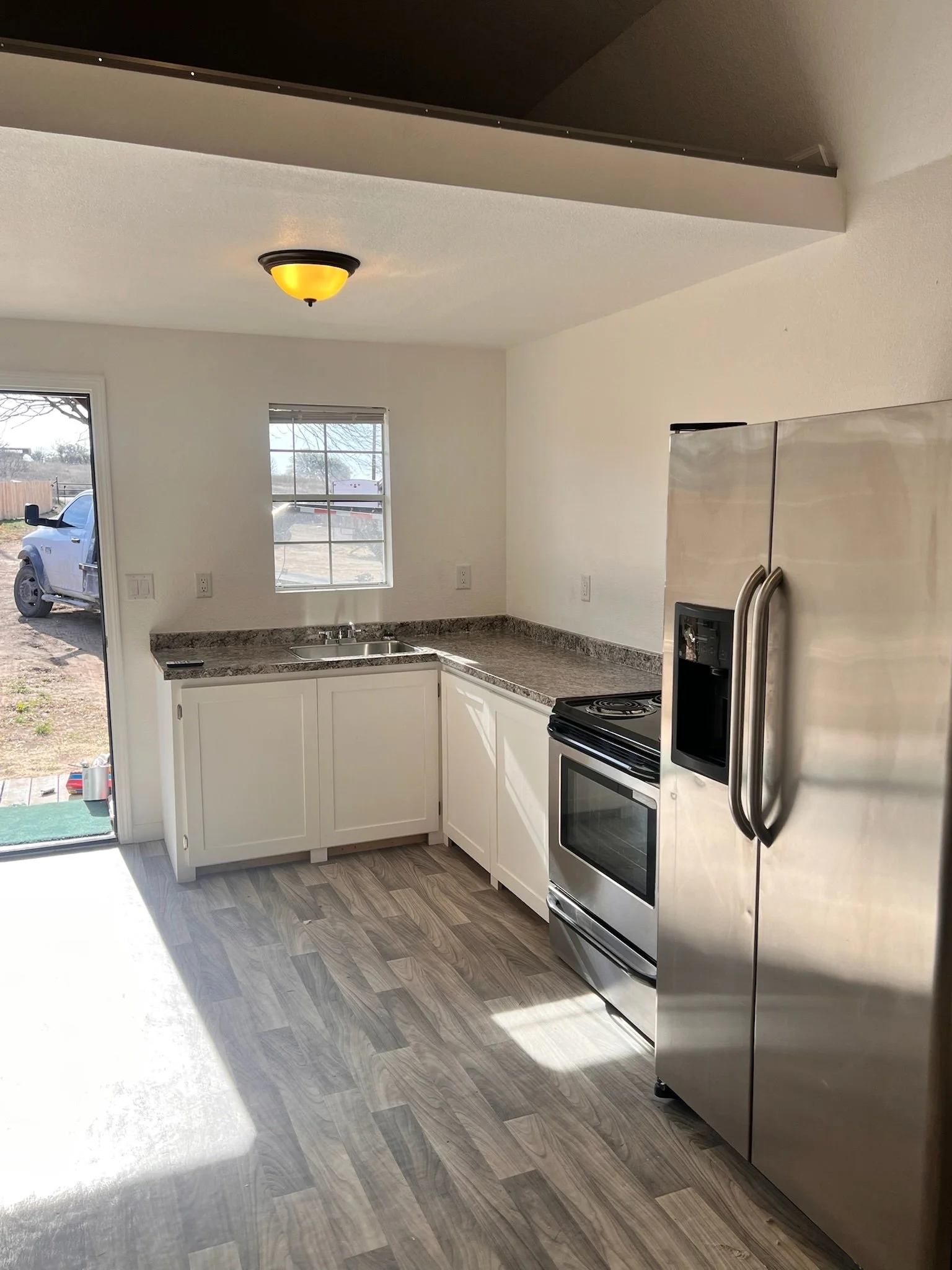 A modern kitchen with white cabinets, a granite countertop, a stainless steel refrigerator, a black and white stove, and two windows offering a view of an outdoor yard with trees and a vehicle. There is a ceiling light fixture turned on.