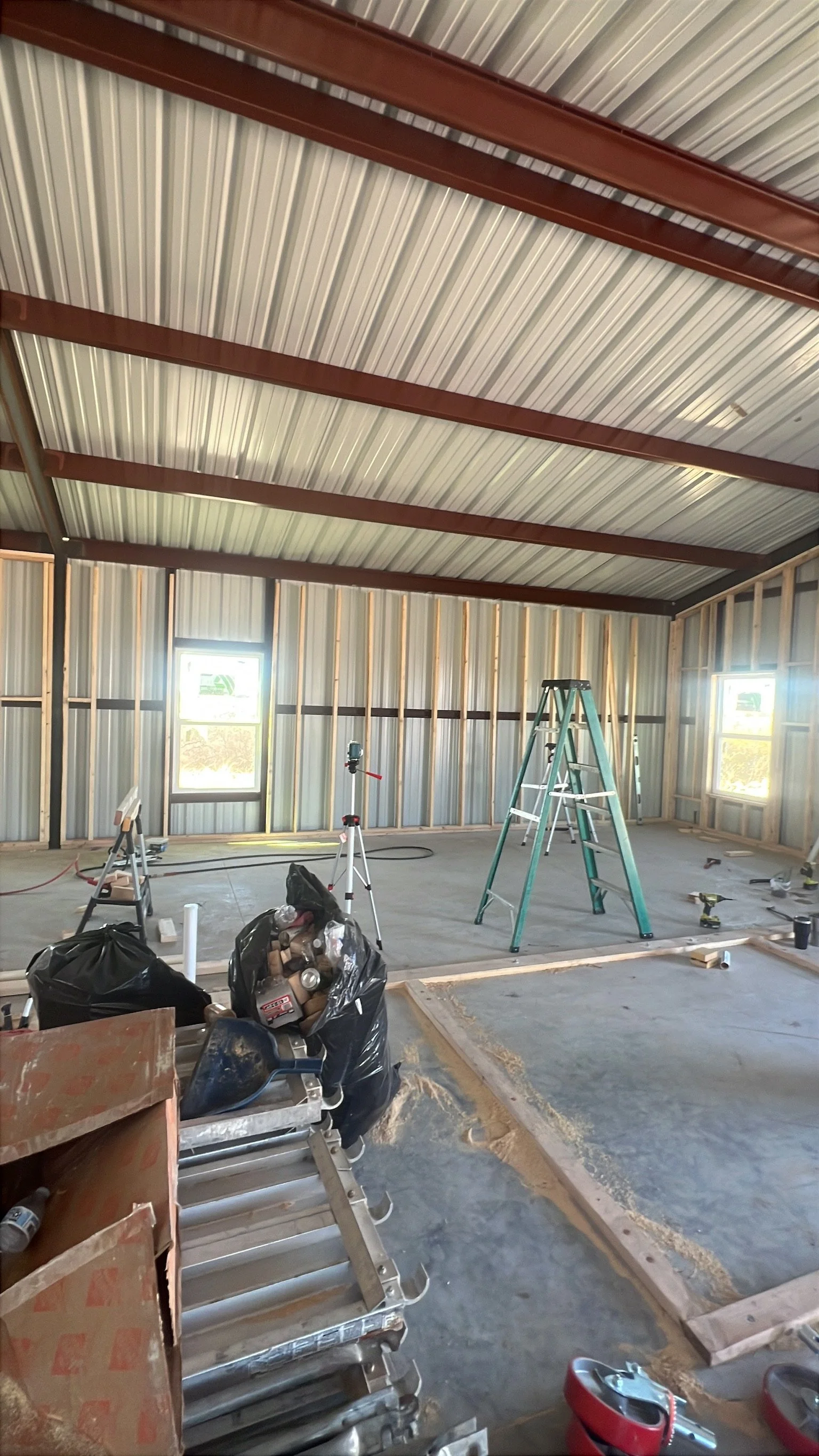 Interior of a building under construction with exposed metal walls and ceiling, construction tools, a ladder, and construction materials scattered around.