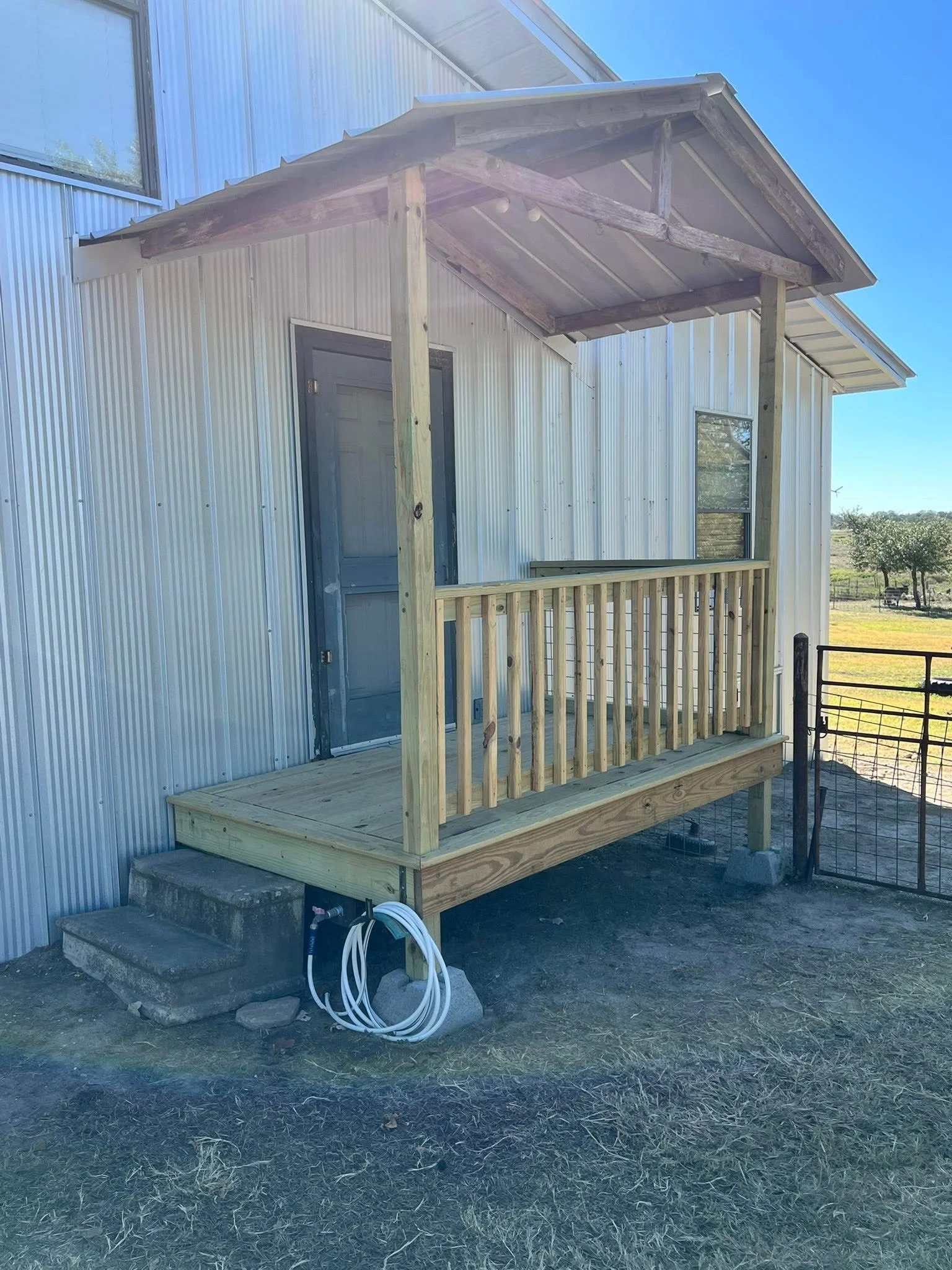 Wooden porch railing and staircase outside a metal building with a door and window, blue sky, and fields in the background.