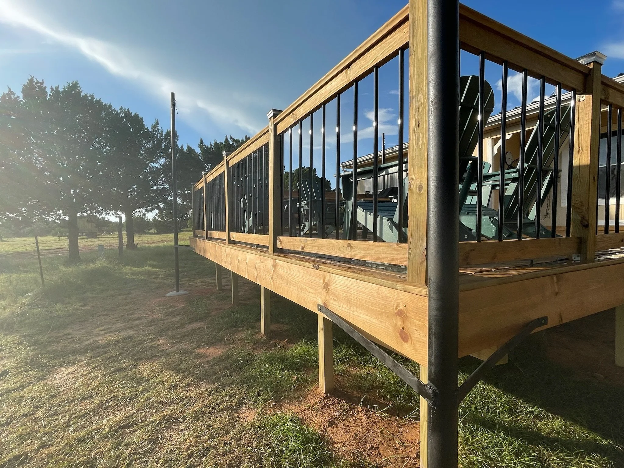 Wooden deck with metal railing, chairs and a satellite dish, in a grassy outdoor area with trees and a blue sky.