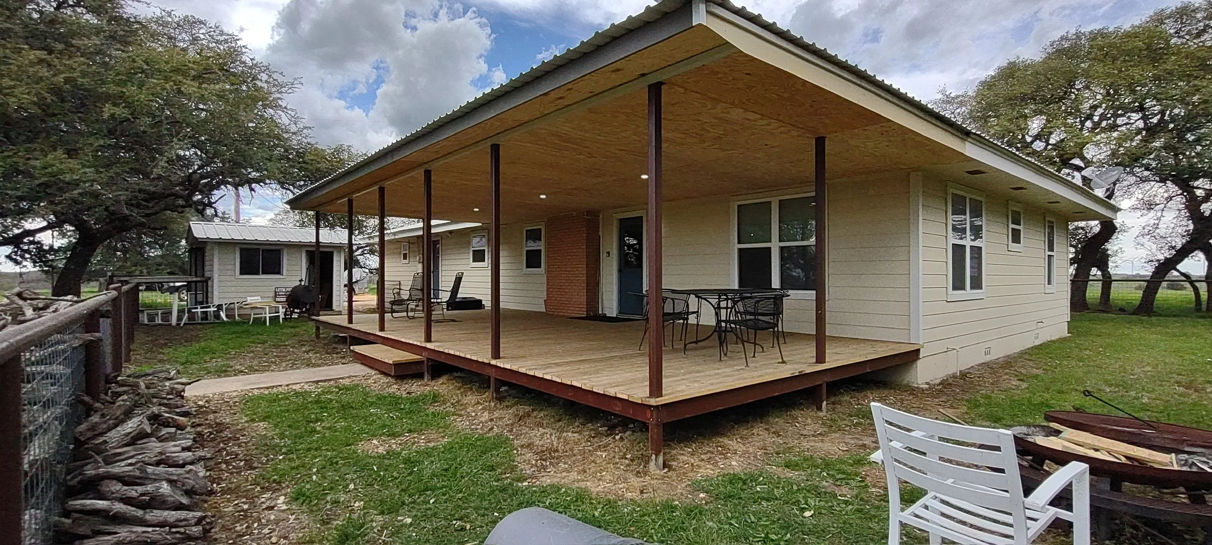 A house with a wooden porch, outdoor furniture, and trees in the background under a partly cloudy sky.