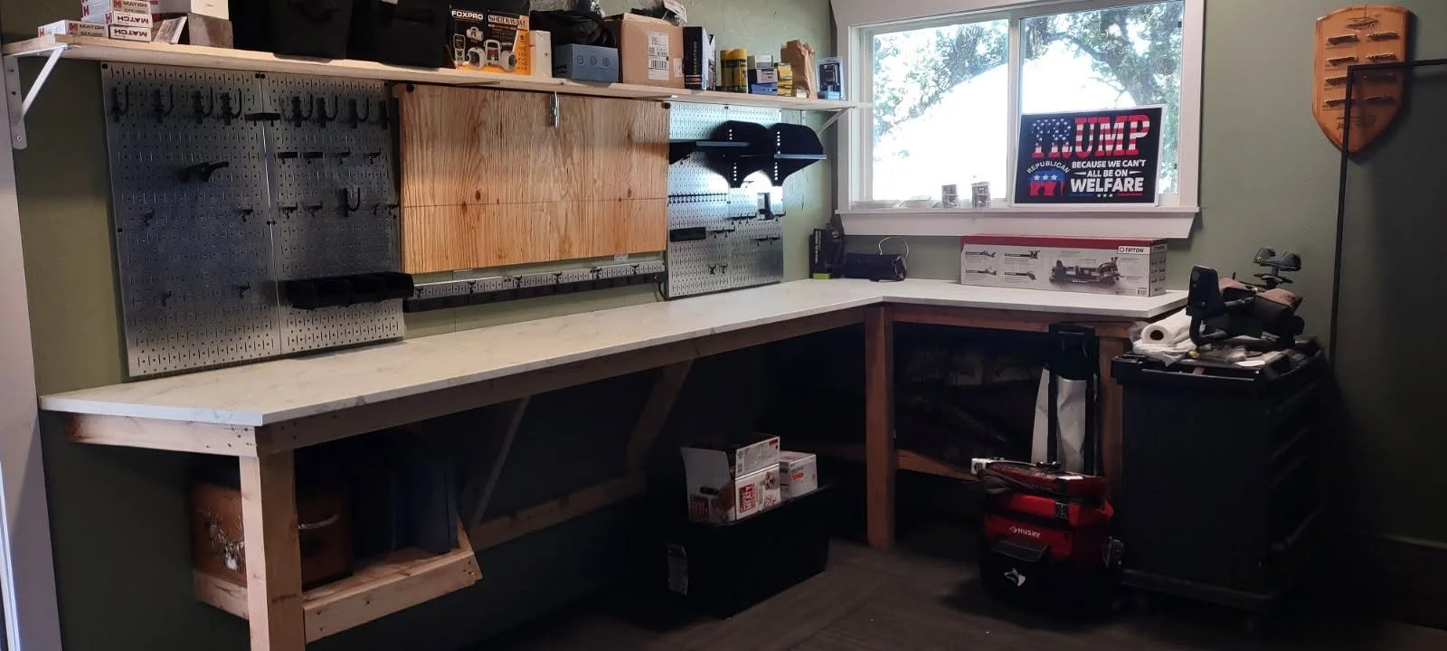 A workshop with a long workbench, pegboard with tools, shelves with boxes, a window, and various equipment and supplies.