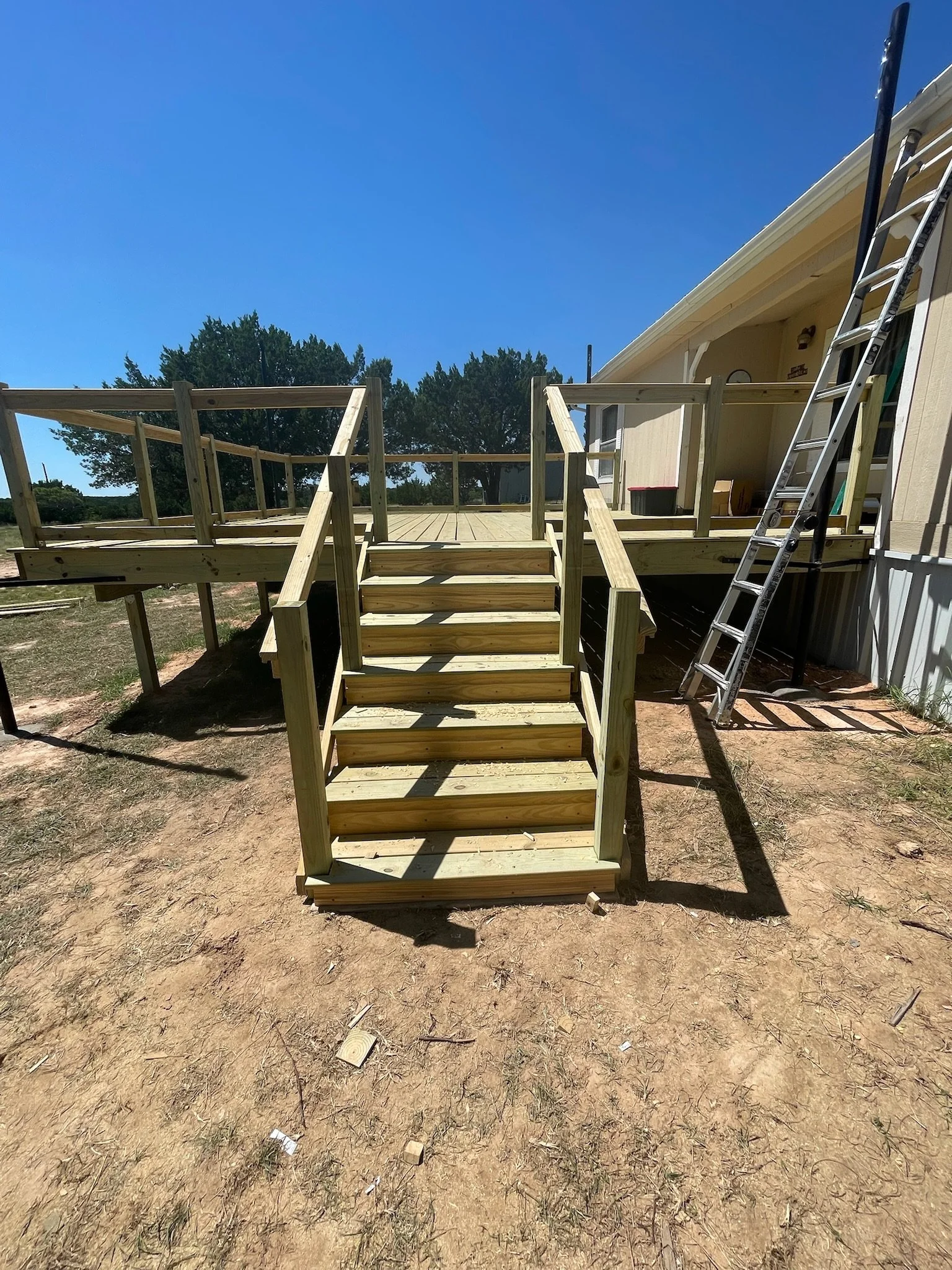 Wooden deck construction with stairs attached to the side of a yellow house, outdoor ladder leaning against the house, and a grassy yard with dirt ground and some small debris.