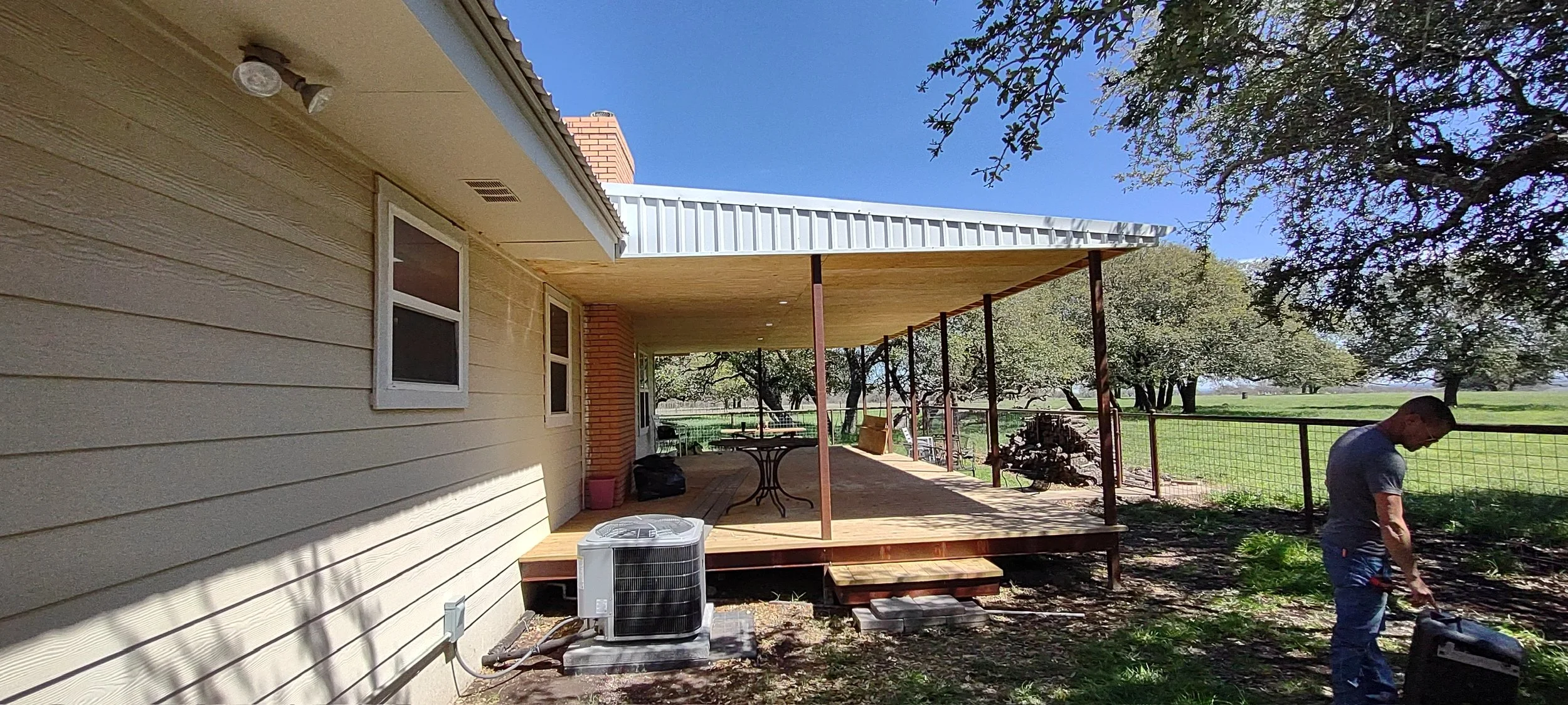 A house with a wooden porch extension being built, with a man working on the ground beside it on a sunny day. The backyard is fenced with green trees and a field in the background.