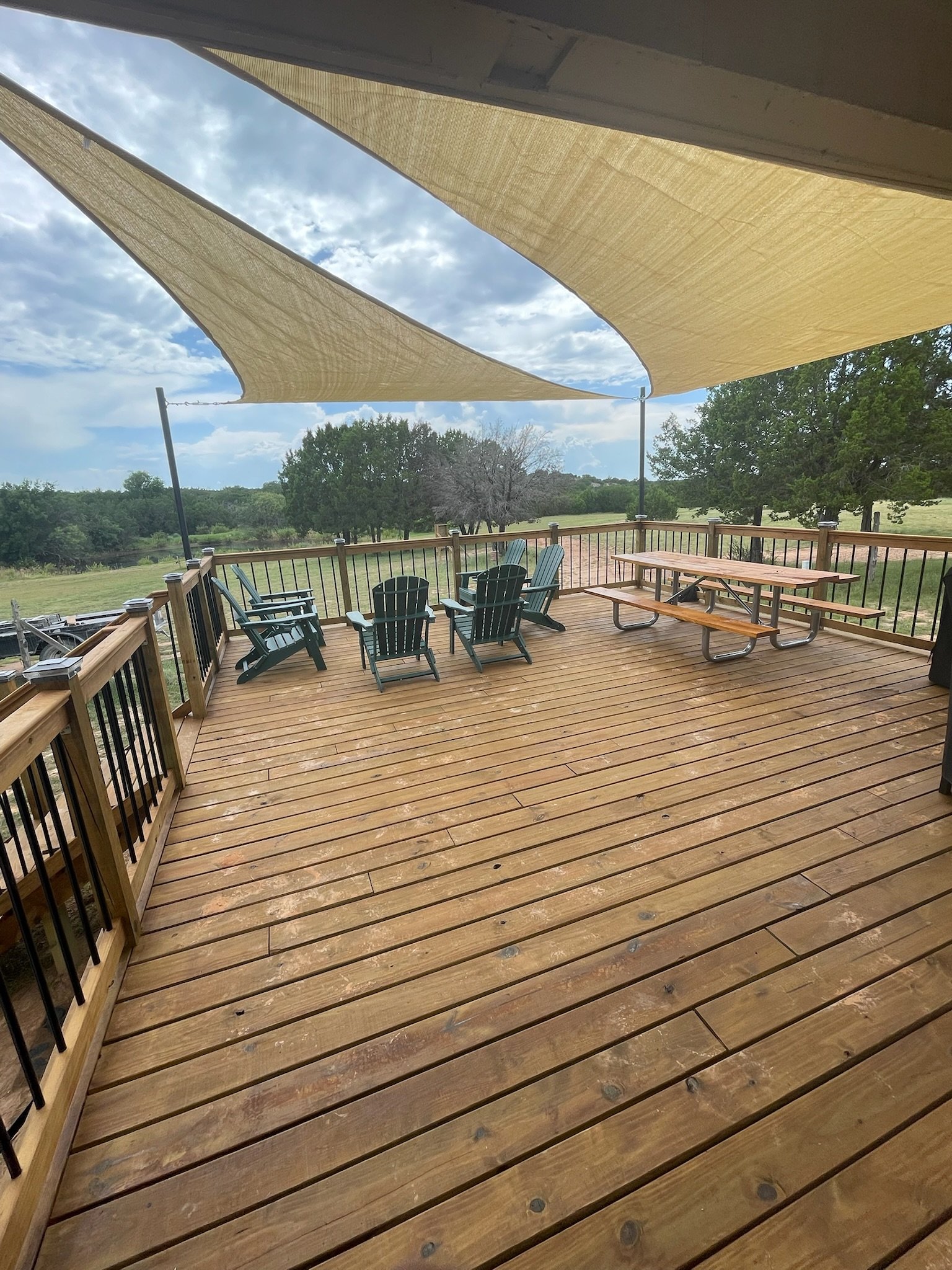 A wooden deck with a railing, outdoor chairs, a picnic table, and shade sails against a backdrop of trees and a partly cloudy sky.