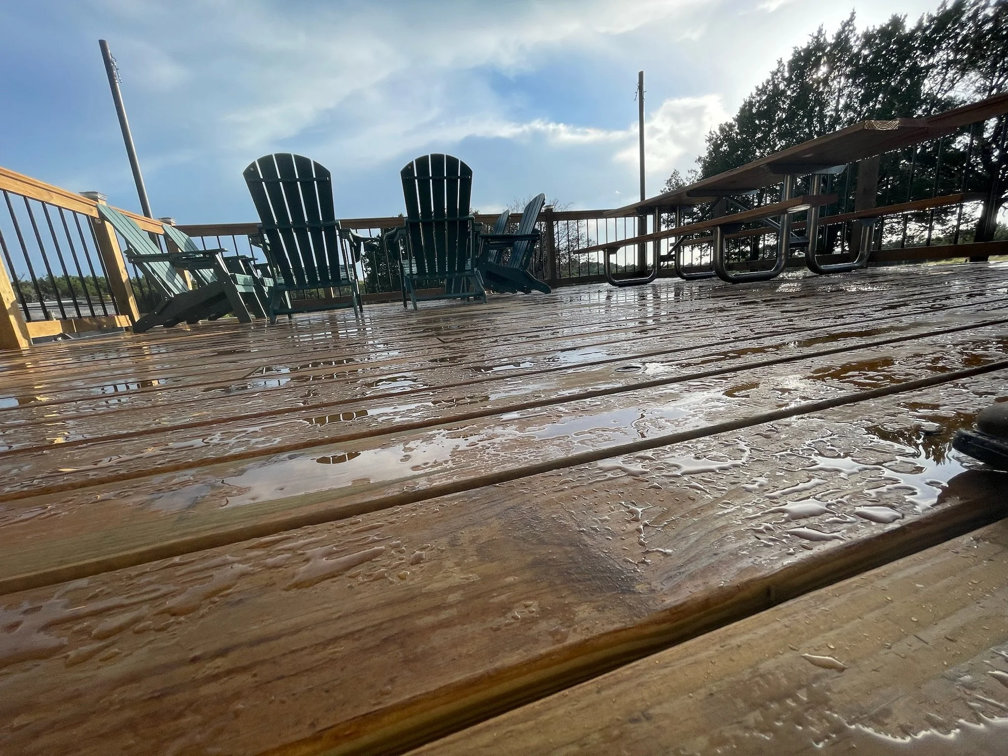 View of a wooden deck with outdoor chairs and tables, wet from rain, with trees and a cloudy sky in the background.
