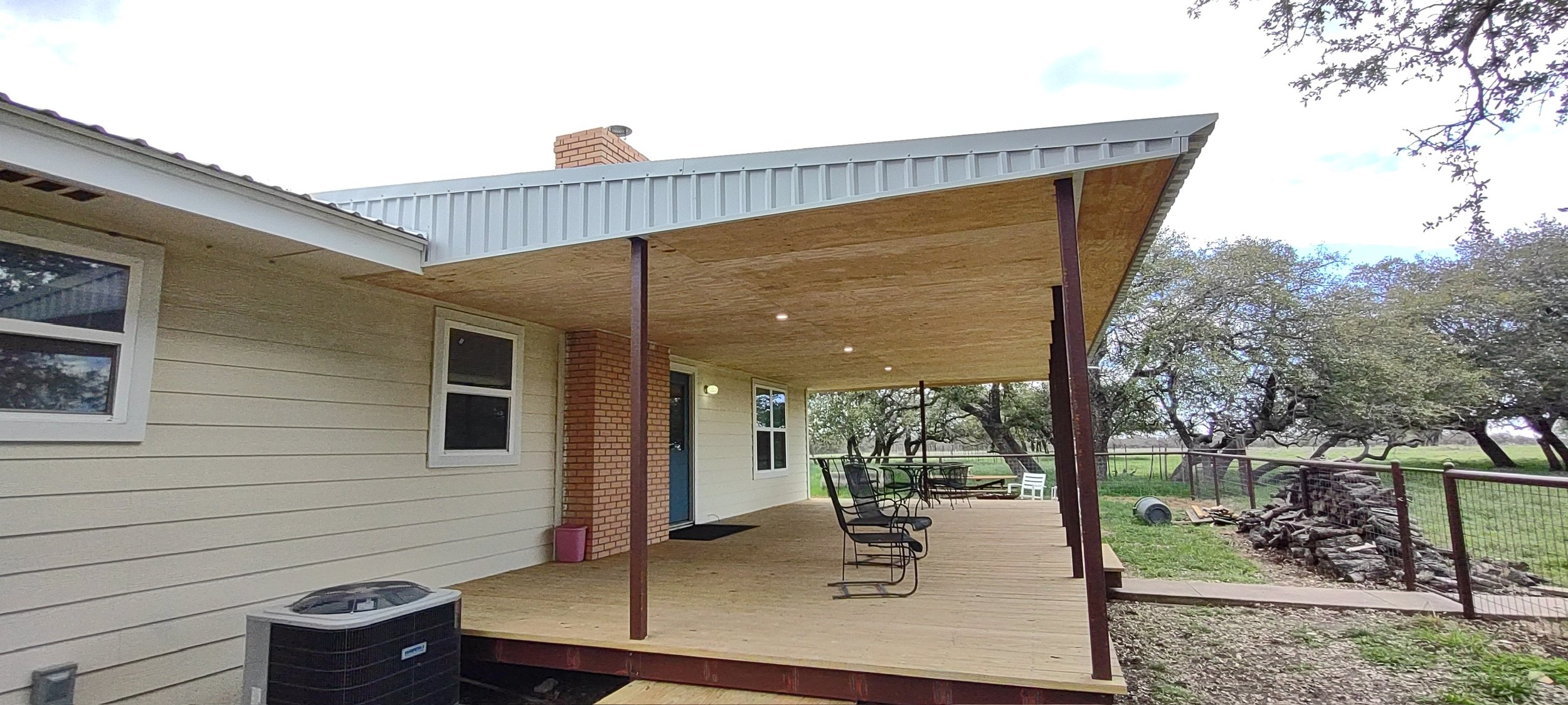 The image shows the outdoor patio of a house with a wooden deck, chairs, and a railing. The house has cream-colored siding, a brick chimney, and multiple windows. There are trees and a fenced yard in the background.
