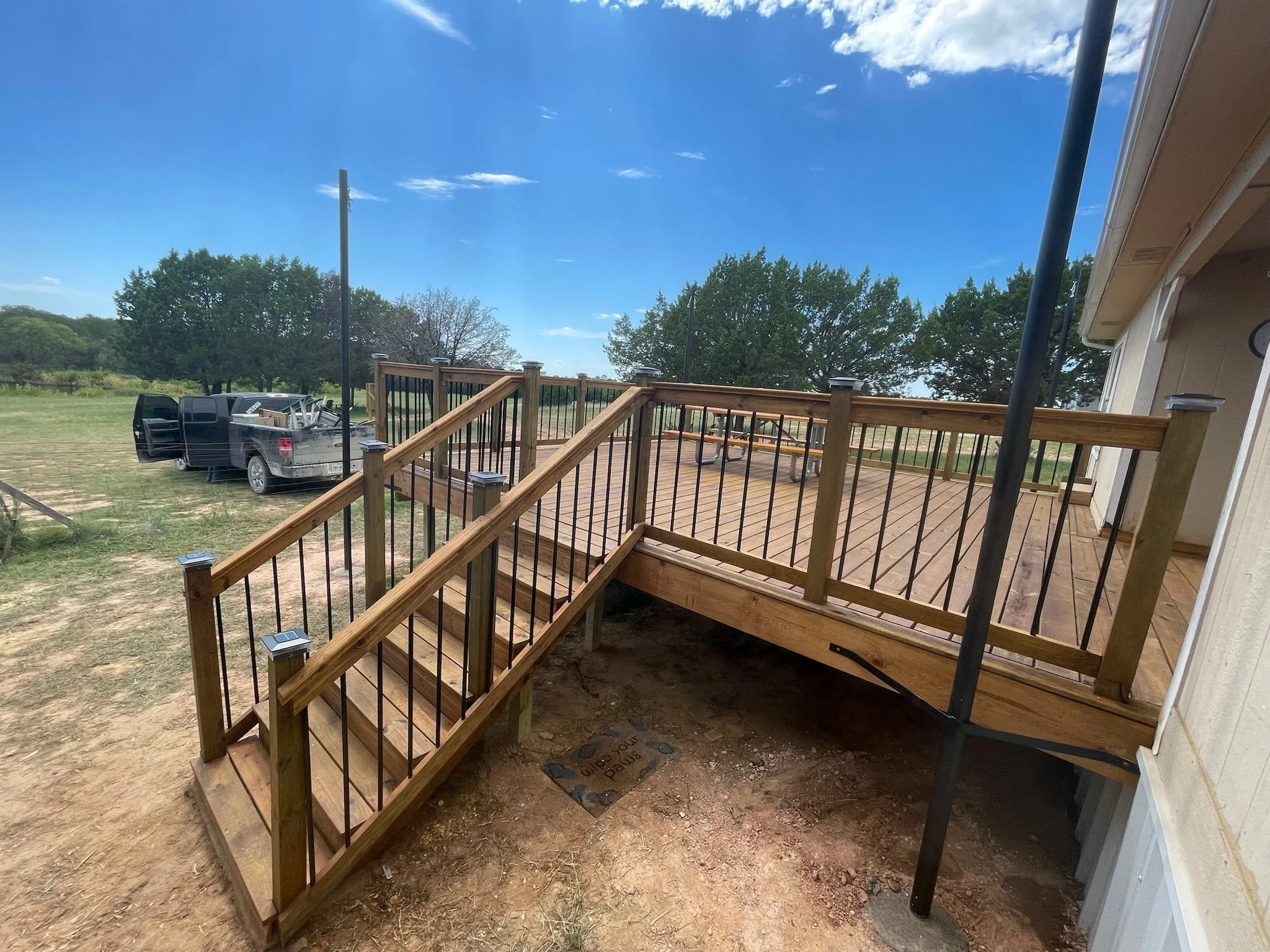 A wooden deck with a railing attached to a house, overlooking a green yard with trees and a black pickup truck. The sky is clear and blue.