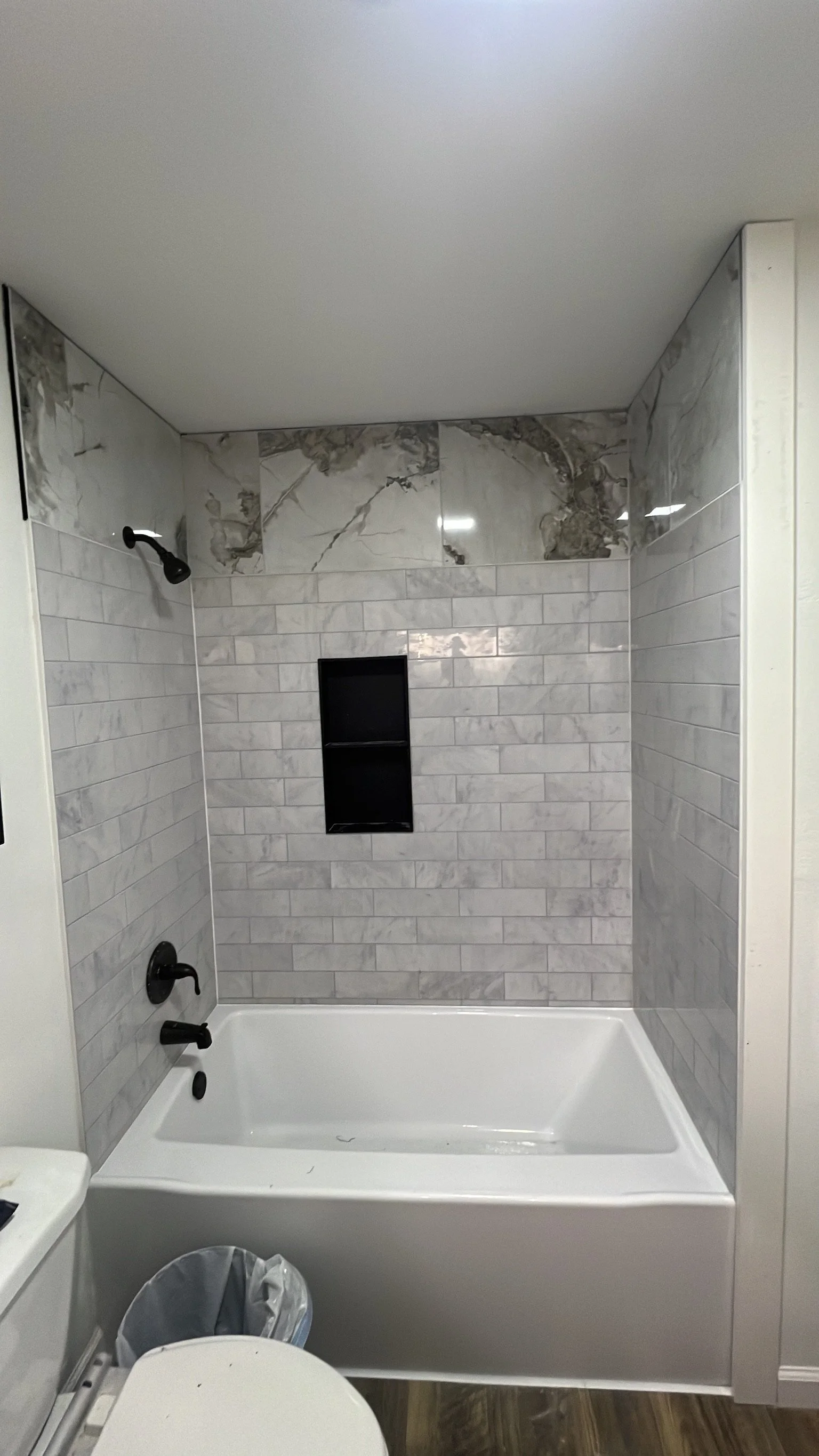 A bathroom shower area with gray and white marble tile walls, a black showerhead, a black shower control handle, a small built-in black shelf, a white bathtub, a trash can, and part of a toilet, with wood-like flooring.