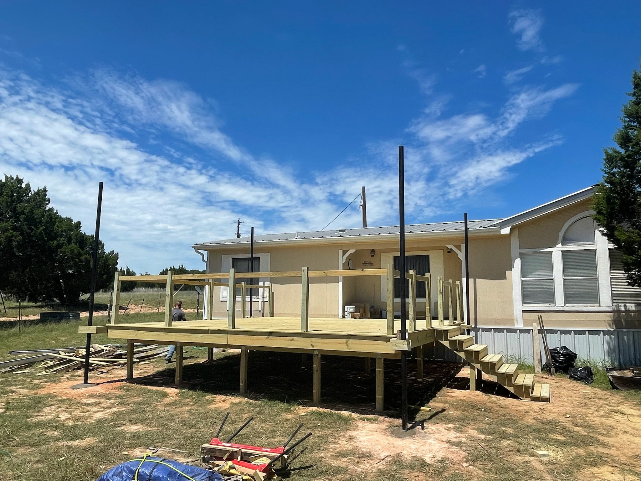 A house under construction with a wooden deck in the backyard, surrounded by green grass and trees. Construction tools and materials are scattered around.