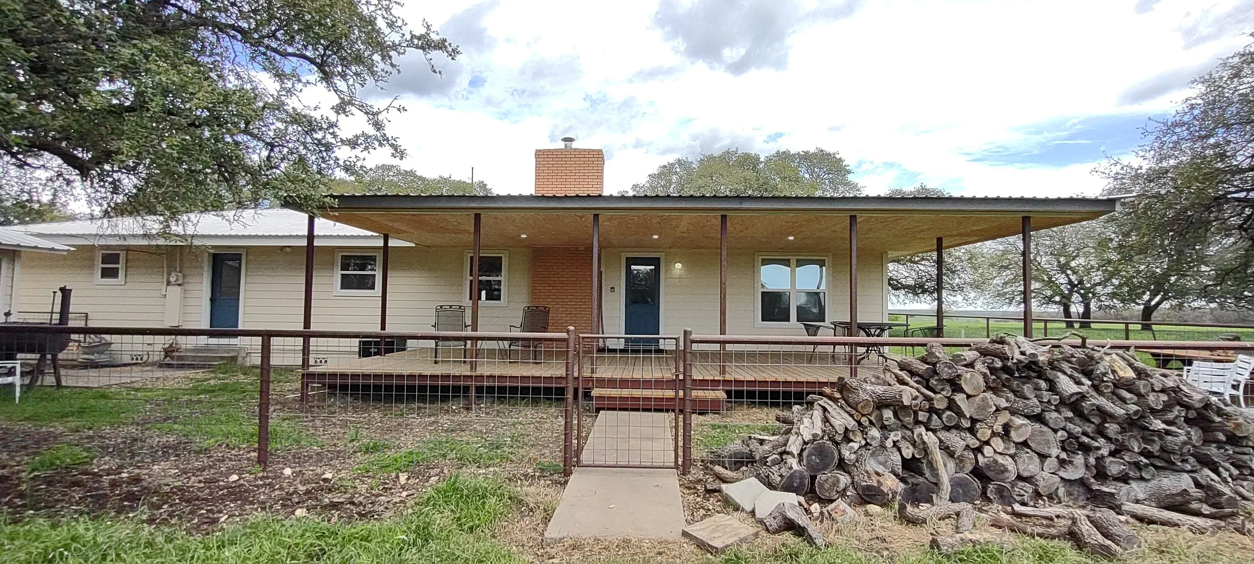 A house with a large covered porch, a brick chimney, and a pile of chopped logs in the front yard. There are trees and a cloudy sky in the background.
