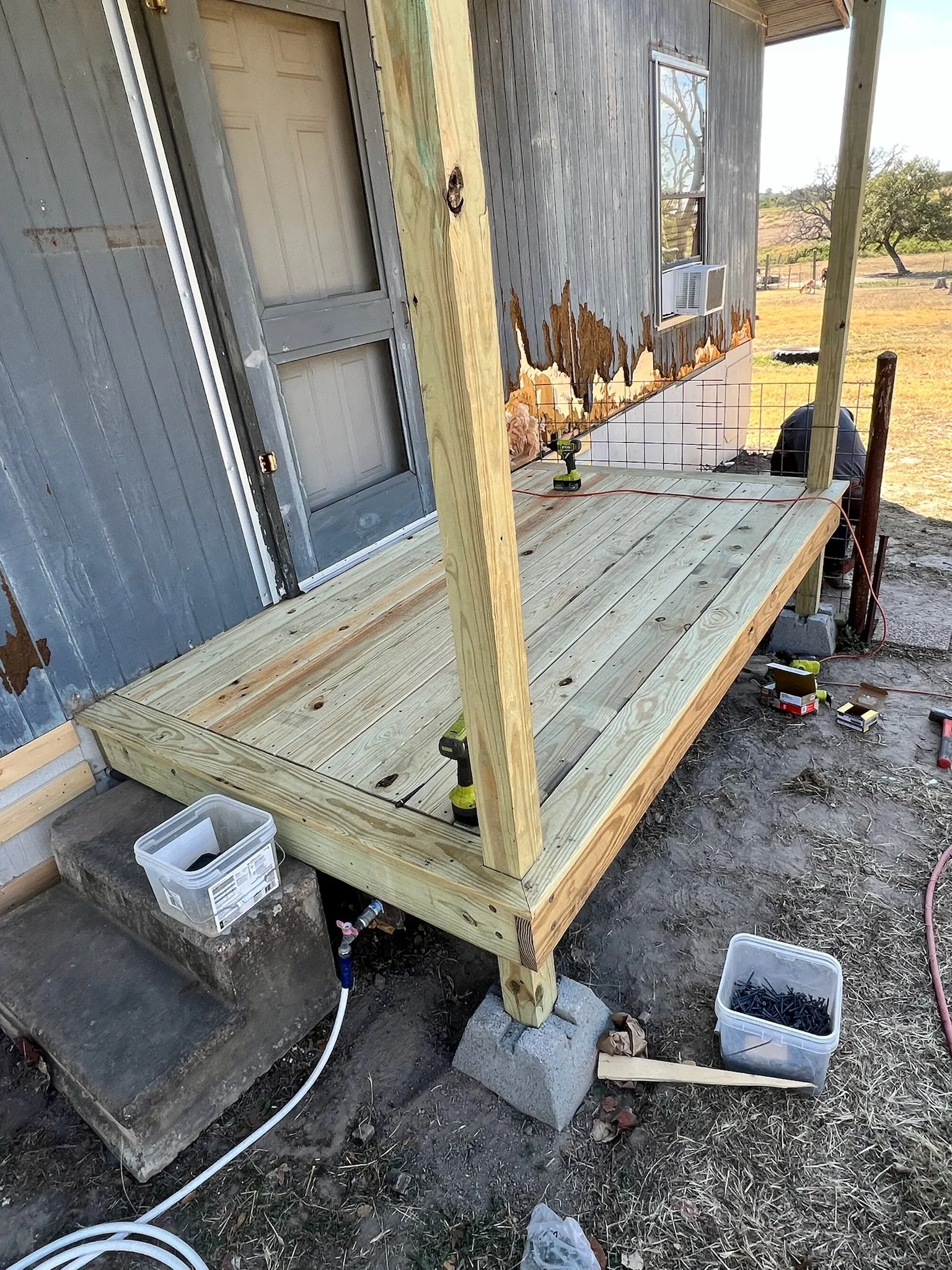 Building a wooden deck attached to a house with gray siding and a window air conditioning unit. Some construction tools and materials are seen around, with new wood framing and support posts in place.