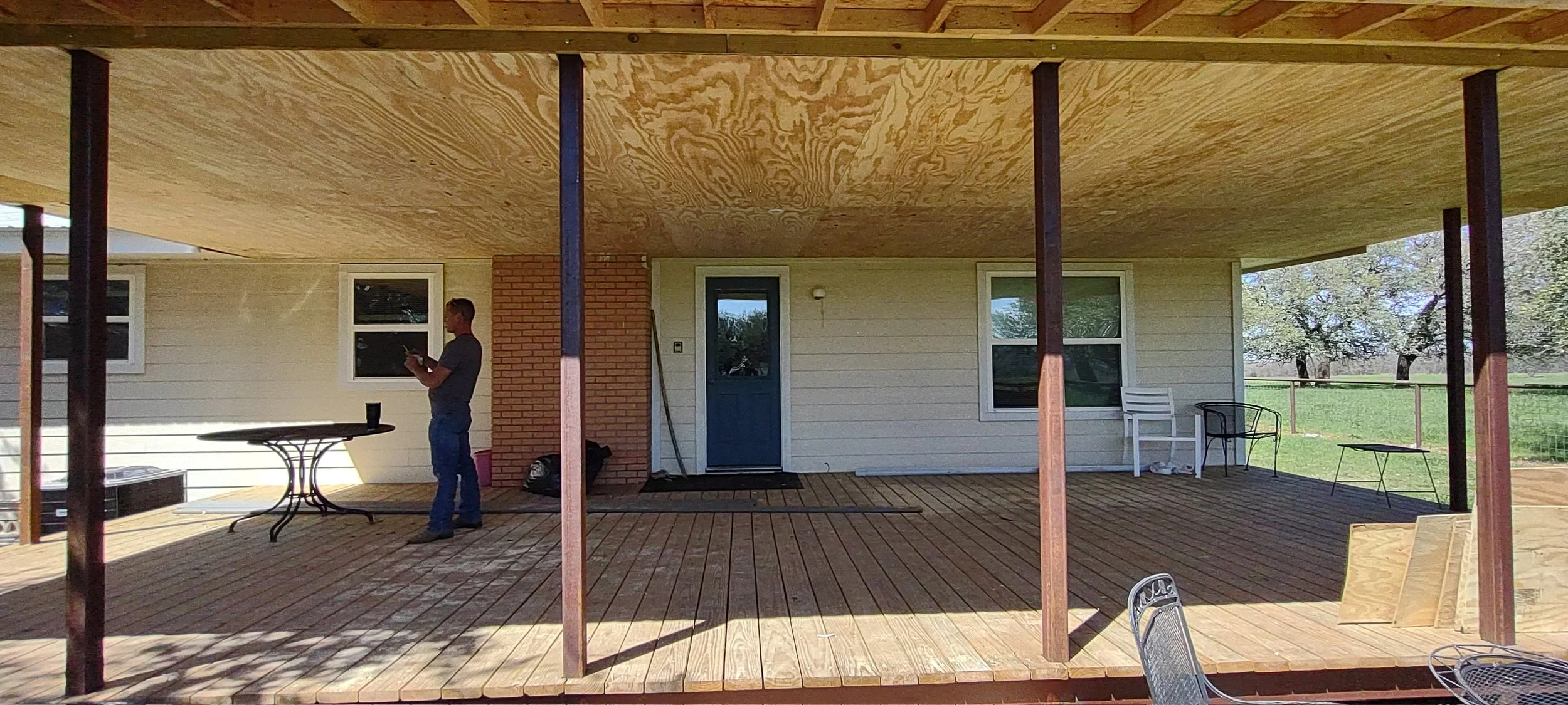 A person standing on a wooden deck under construction, with a house in the background. The house has white siding, a blue door, and multiple windows. The deck has several chairs and a table, and the underside of the roof is unfinished plywood.