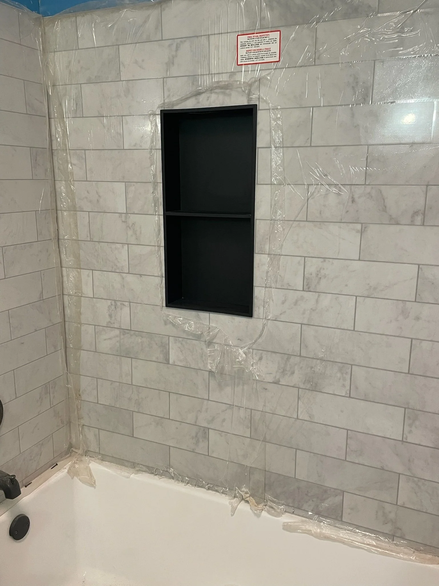 Bathroom wall with white marble tiles, a black wall shelf, and a red and white inspection notice at the top. Clear plastic protective covering is taped around the tiles and shelf.