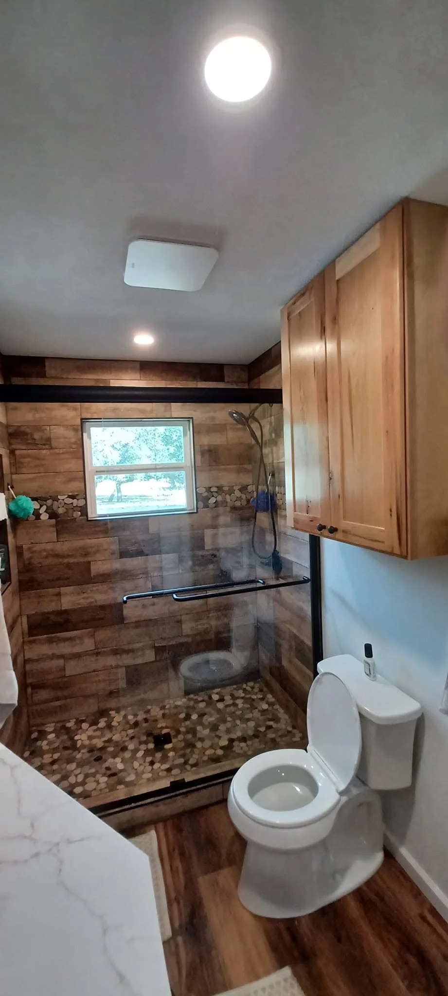 Bathroom with a walk-in shower featuring wood paneling, pebble flooring, and a small window, alongside a toilet and wooden cabinets.