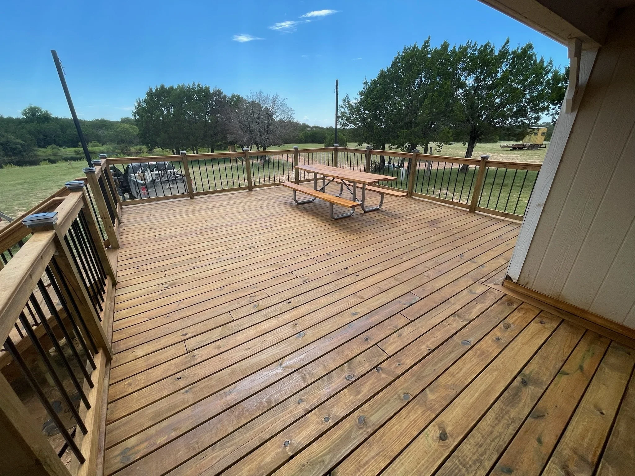 A wooden deck with a picnic table and benches, overlooking a grassy field with a few trees and a pond, under a blue sky with some clouds.