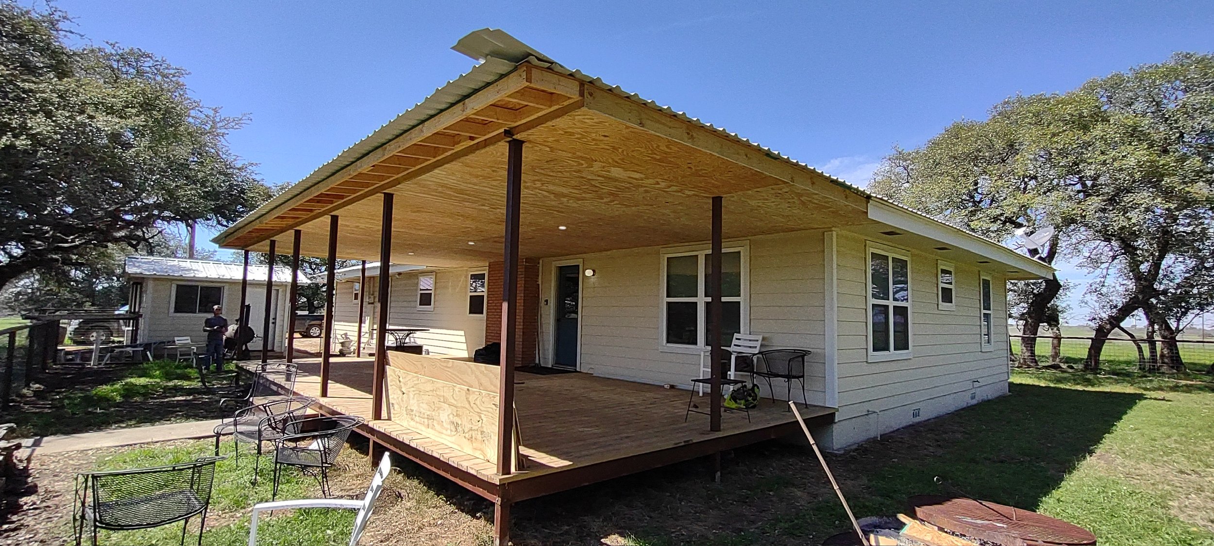 A house with a deck under construction, with a person in a black shirt and cap standing outside, surrounded by trees and lawn.