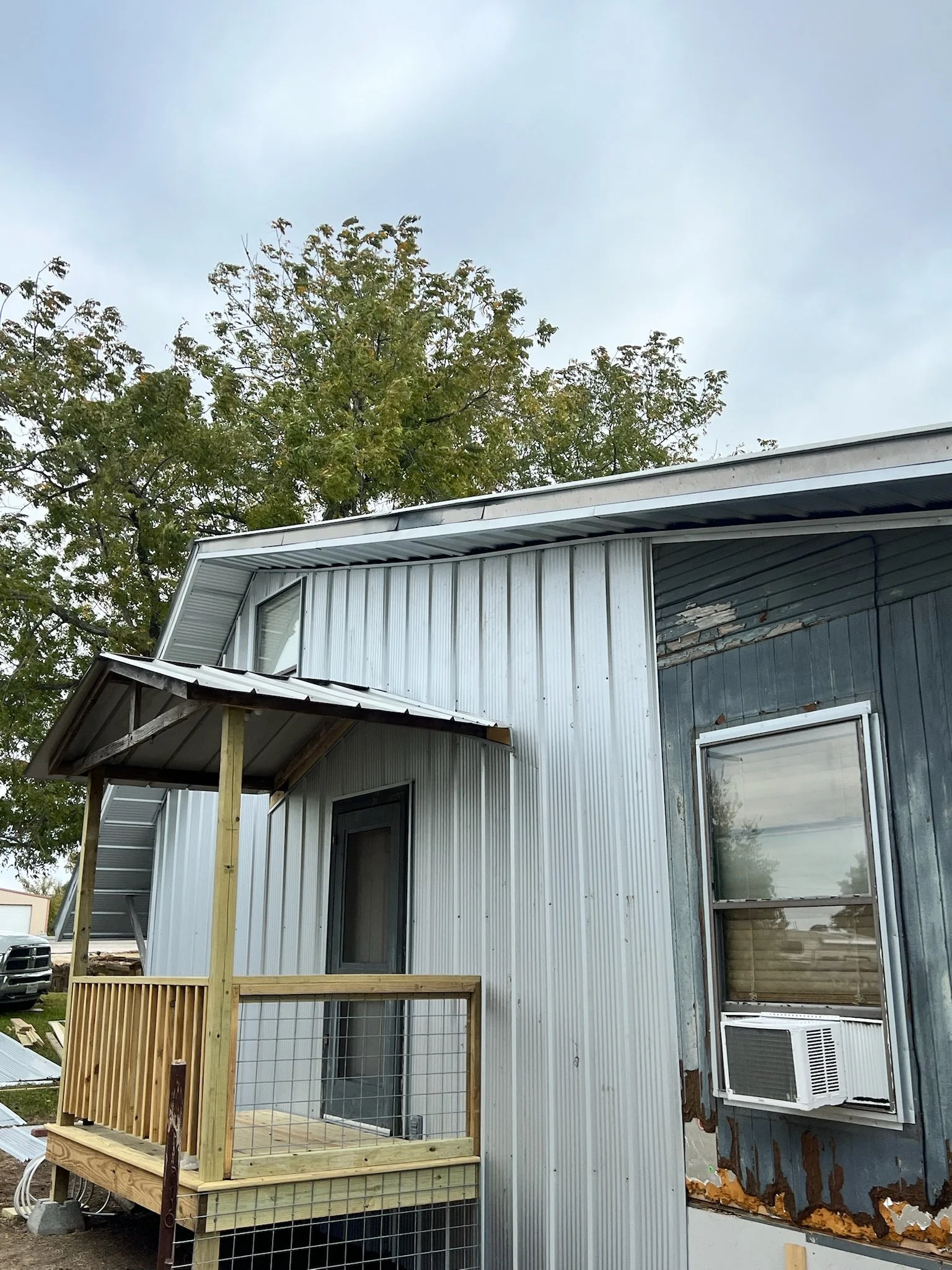 A house with metal siding, a small screened porch with wooden framing under construction, and an air conditioning unit in the window. There is a large tree in the background under a cloudy sky.