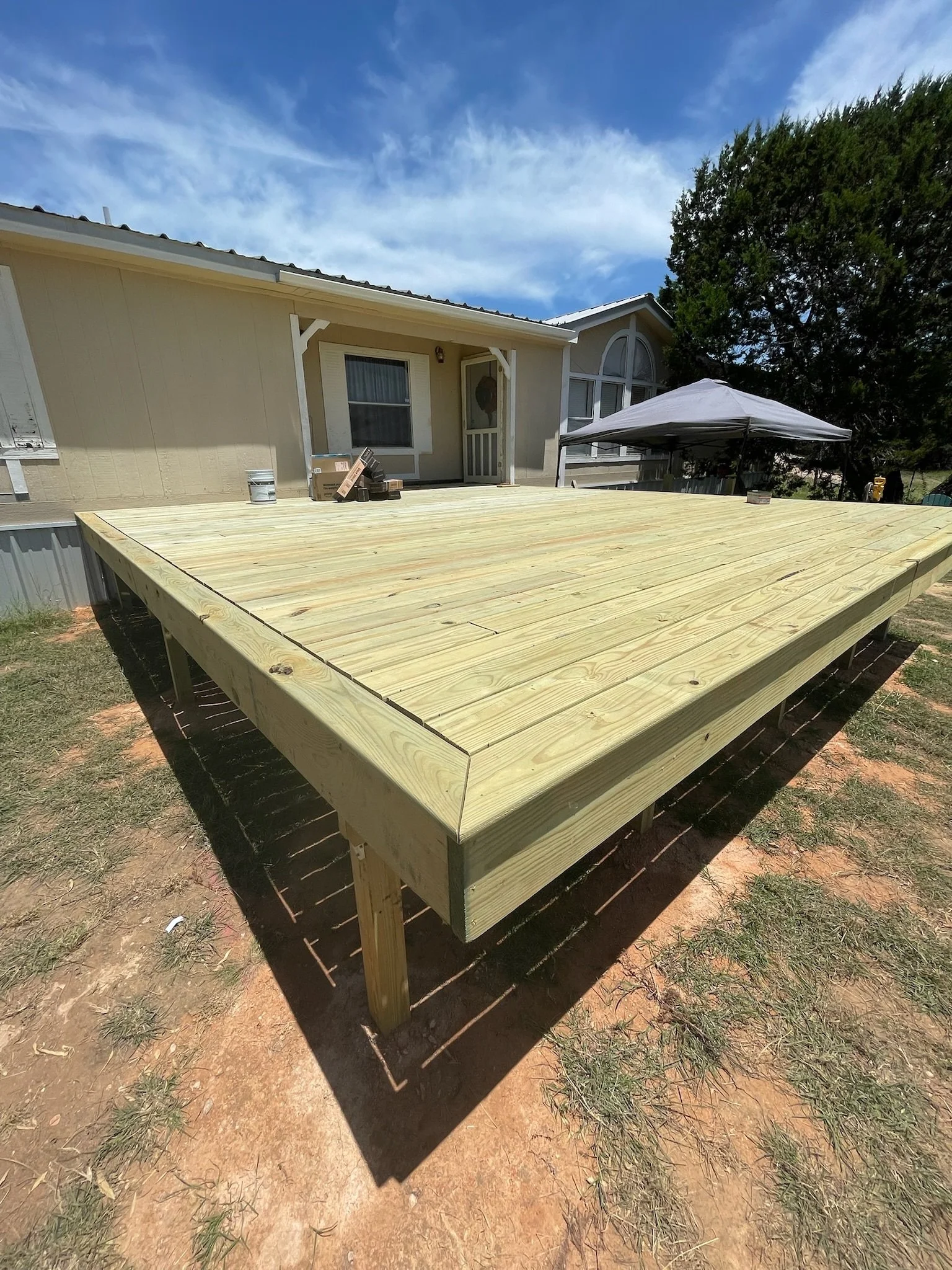 Newly built wooden deck in backyard with tools and supplies for construction. House with beige siding, window, and patio door in background. Large trees and a blue sky with clouds.