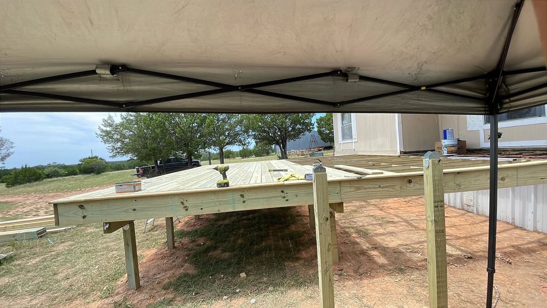 Under a canopy, a construction site shows a wooden deck being built outside a house, with tools and materials on the deck, trees and a truck in the background.