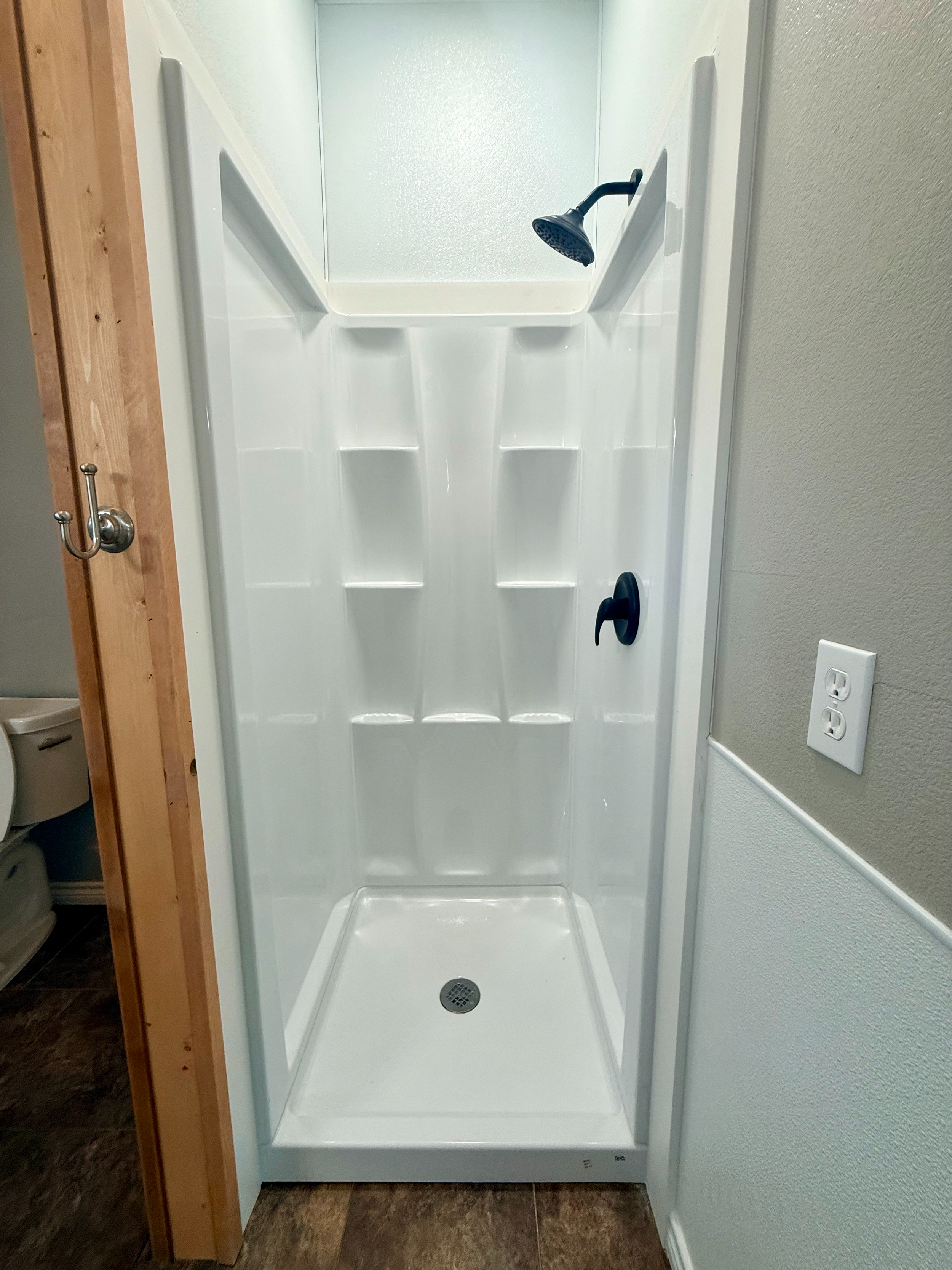 White shower stall with a black showerhead, a drain at the bottom, and wall shelves in the back. The stall is situated in a room with a brown tiled floor and a power outlet on the right side.