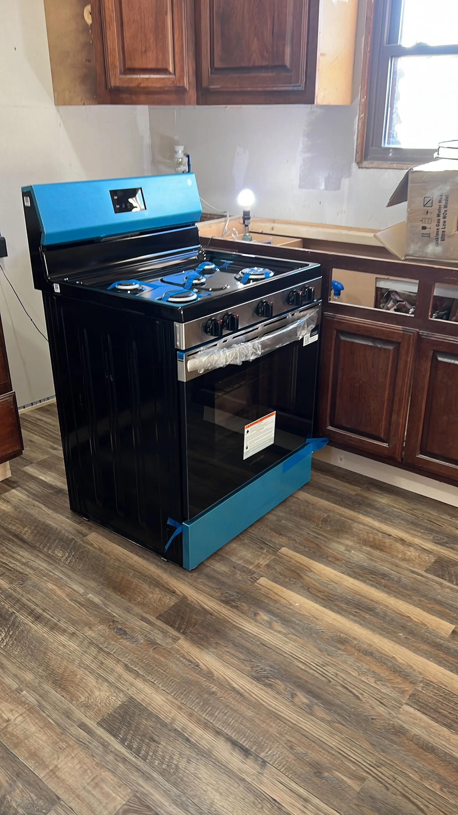 A black and blue gas stove with four burners in a kitchen under renovation. Wood cabinets and a window are visible, with some construction materials and a small lamp nearby.