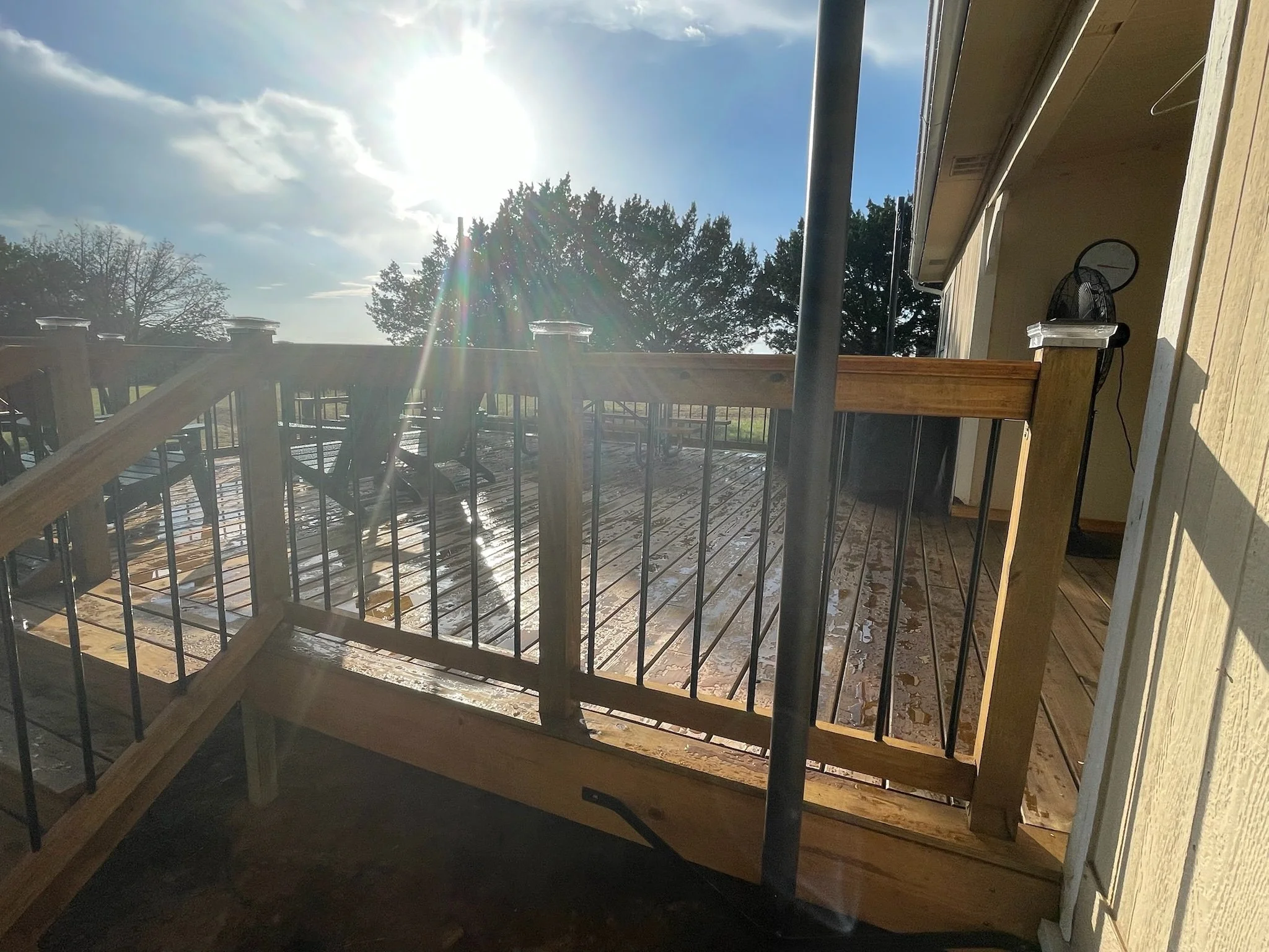 View from a balcony or deck showing a wooden railing, a tree, a partly cloudy sky with the sun shining brightly, and a portion of a house with an outdoor fan on the right side.