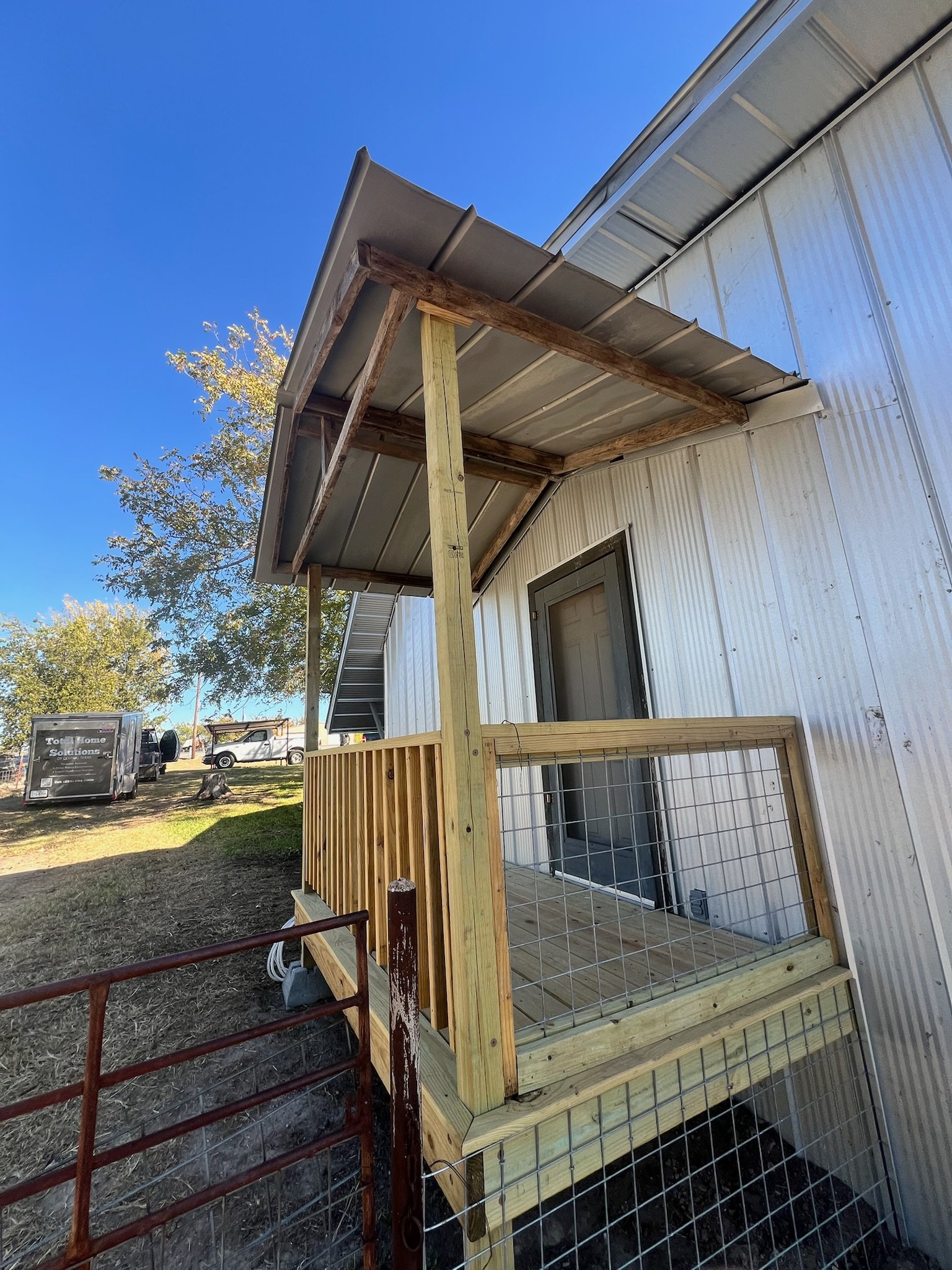 Wooden deck under construction outside a metal building with a sliding glass door, with trees and a clear blue sky in the background.