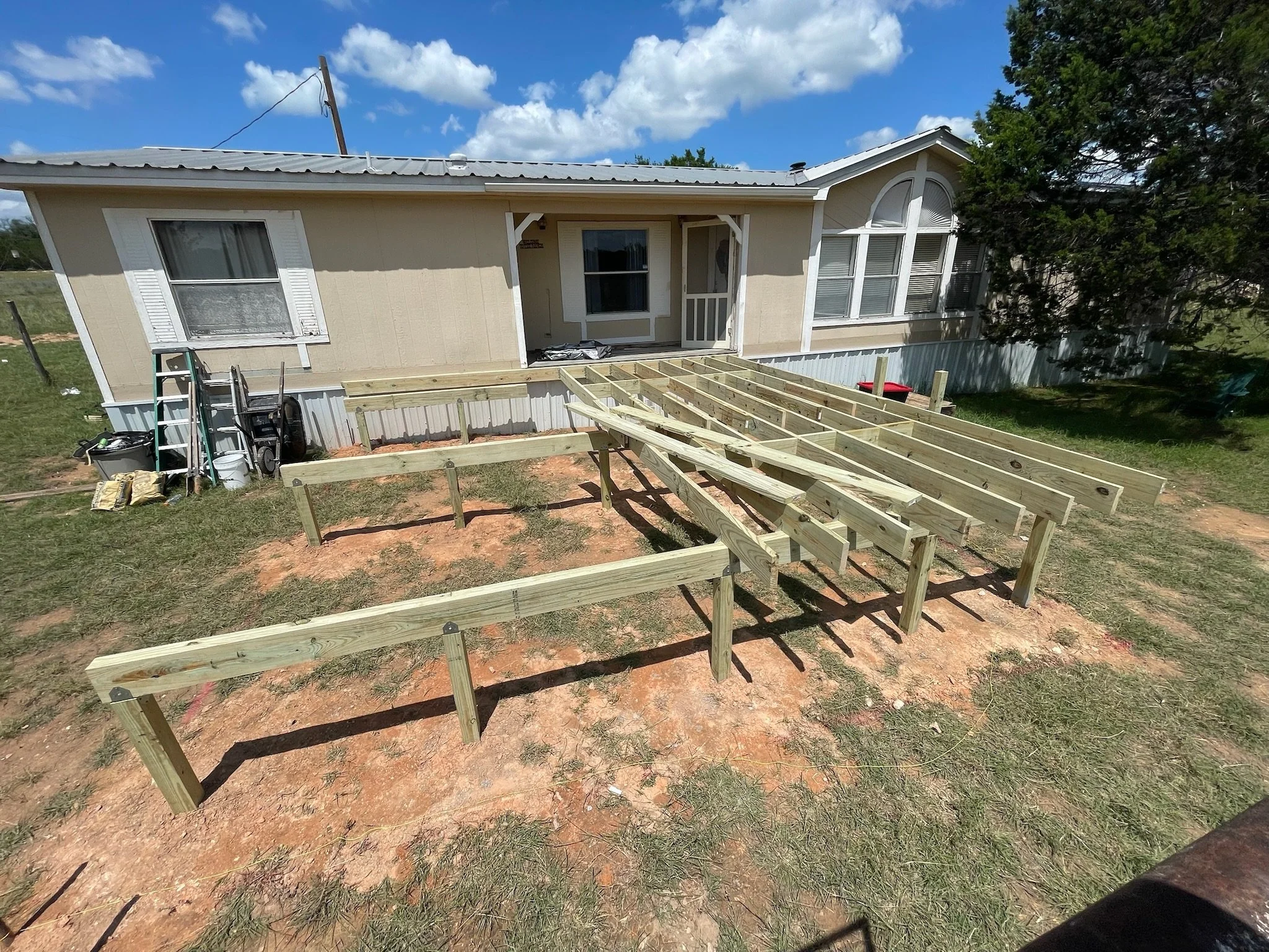 A house under construction shows a front porch with a wooden frame for a deck. There are various construction tools and materials around the area. The house has beige siding, a front door, and a large window, with a tree on the right side and clouds 