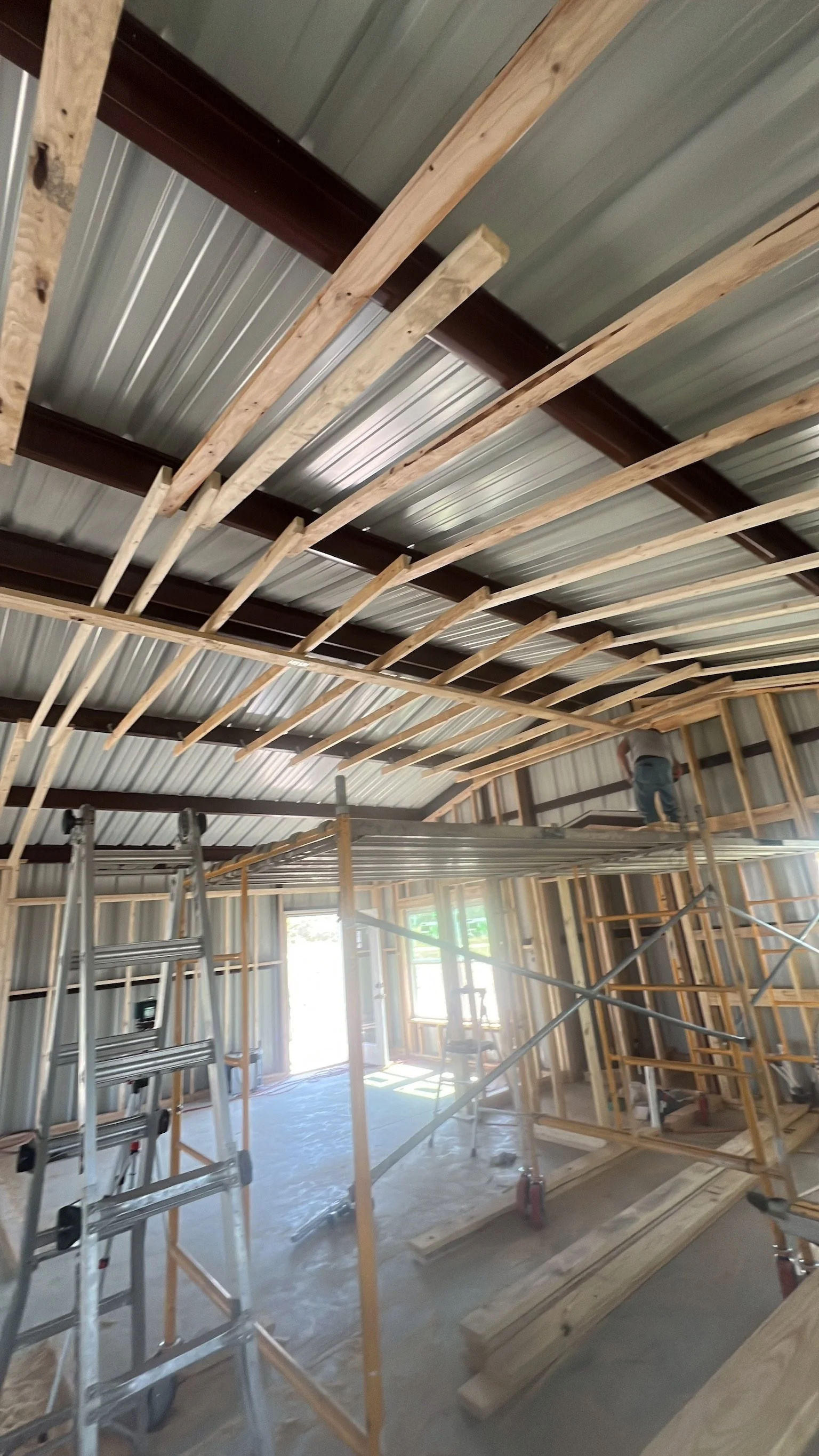 Interior of a building under construction, showing exposed wooden framing, metal roofing, scaffolding, and ladders; a worker is visible in the background.