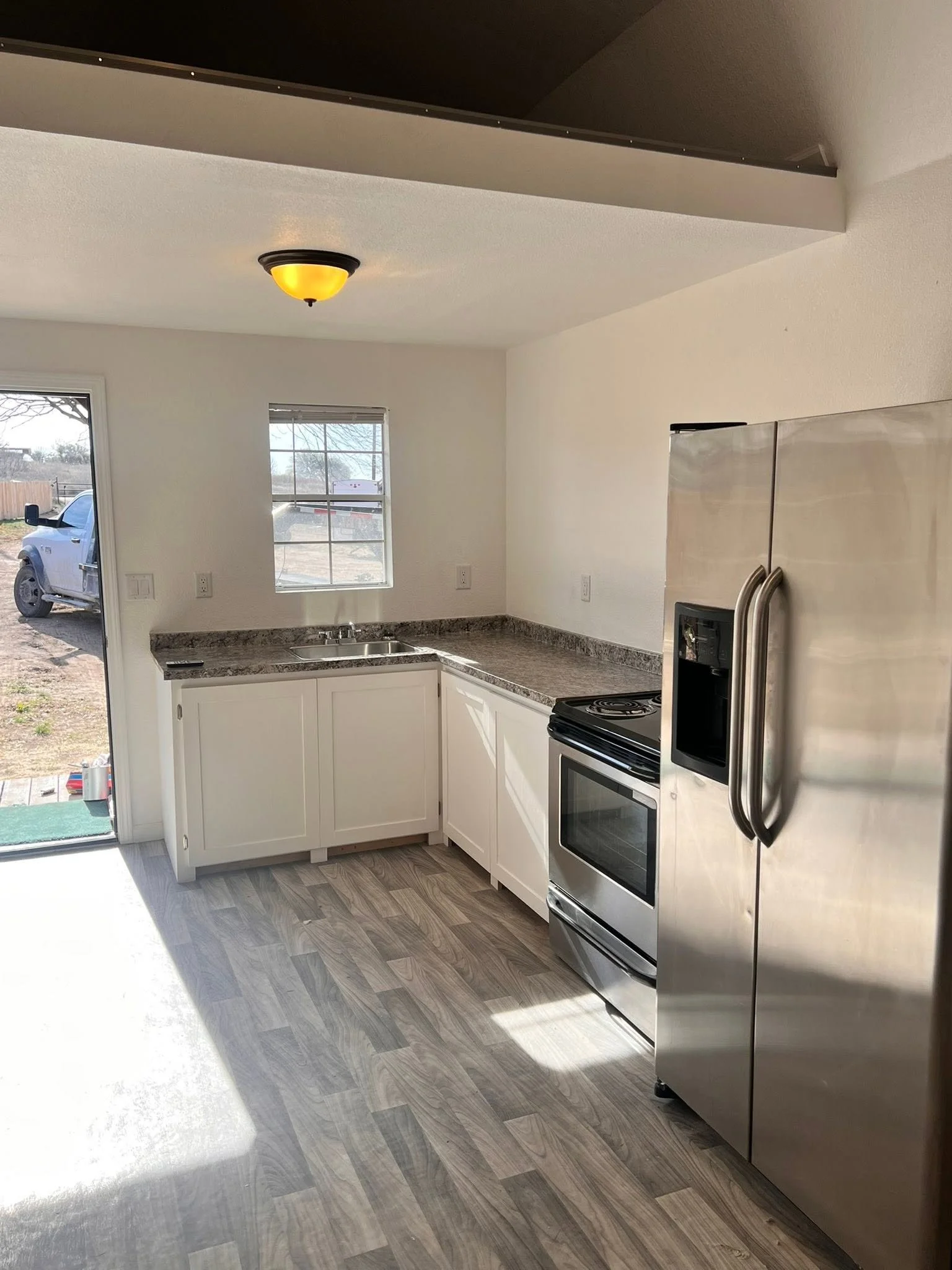 Modern kitchen with white cabinets, a stainless steel refrigerator, a black electric stove, and a window showing an outside yard and a parked vehicle.