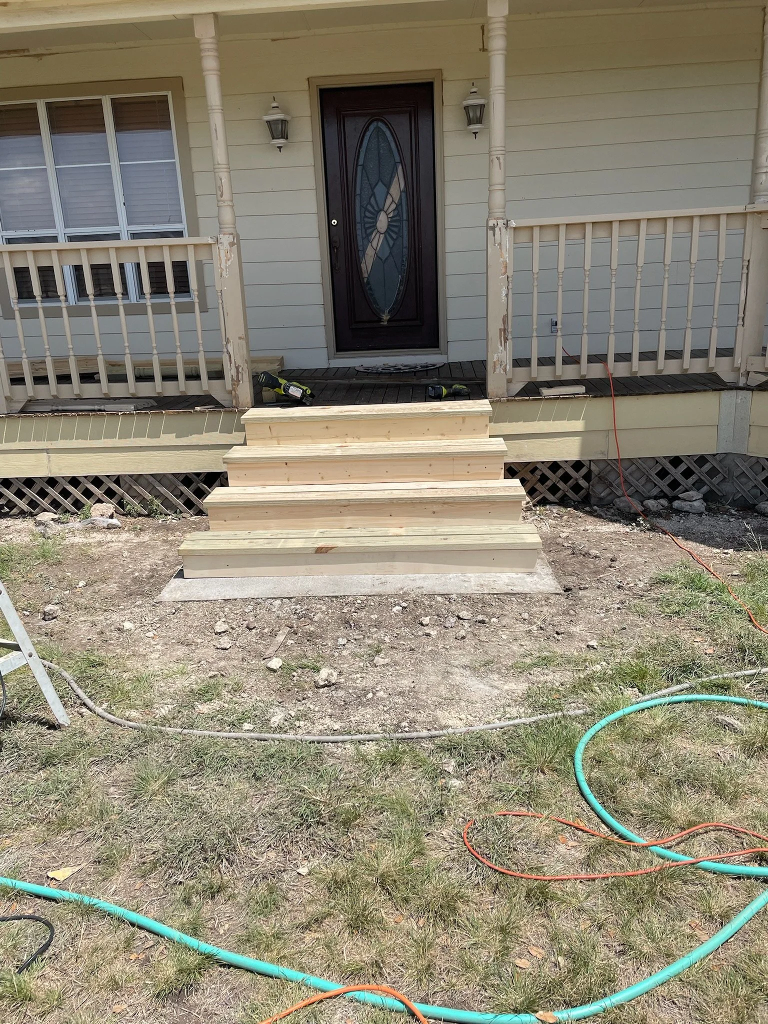 Front porch of a house under construction or renovation. Wooden steps lead up to a front door with tools and hoses on the ground, and the porch railing appears to be in the process of being painted or repaired.