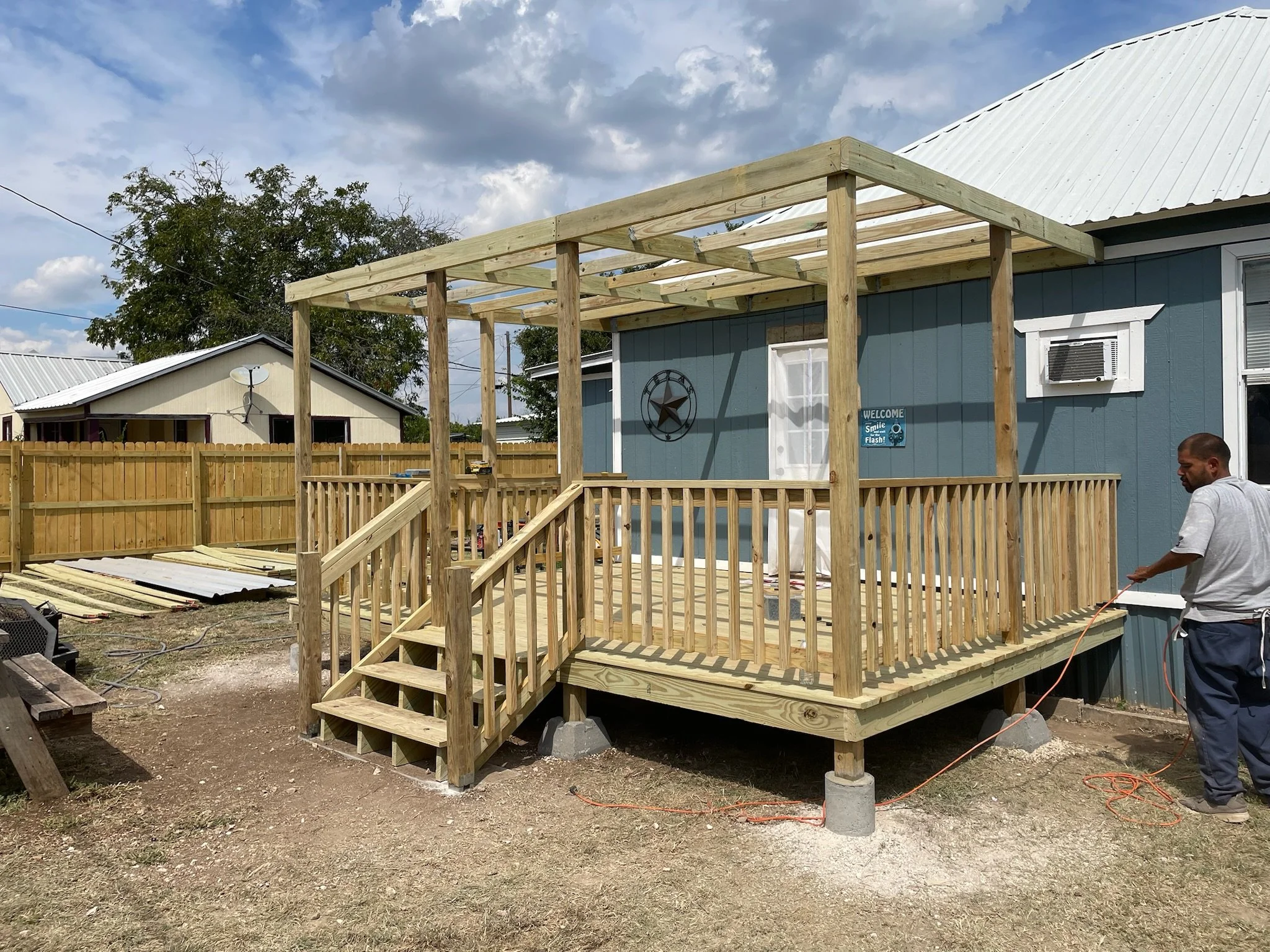 The photo shows a wooden deck under construction attached to a blue house with a white roof. A person is working on the deck, and another person is visible on the right side of the image. The area is outdoors with a wooden fence and other houses in t