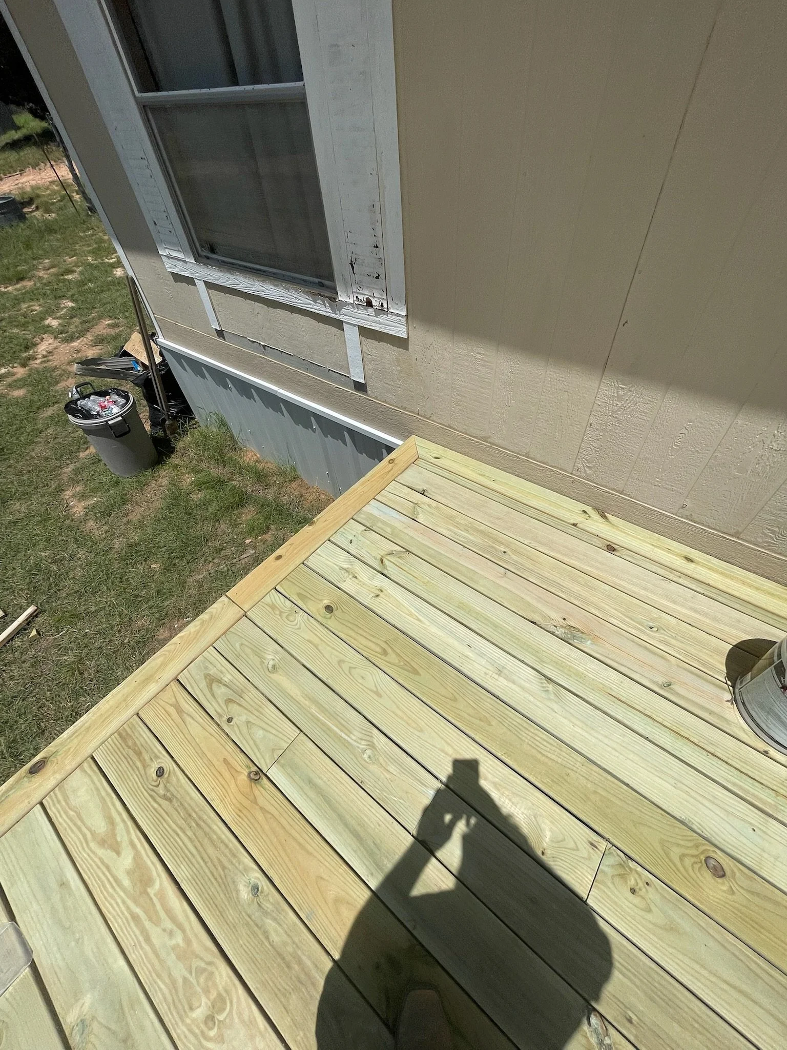 Photo of a wooden deck with new light-colored wooden planks. In the background, there is a house wall with a window and a grass yard with an outdoor trash can and some scattered items. A person's shadow is visible on the deck.