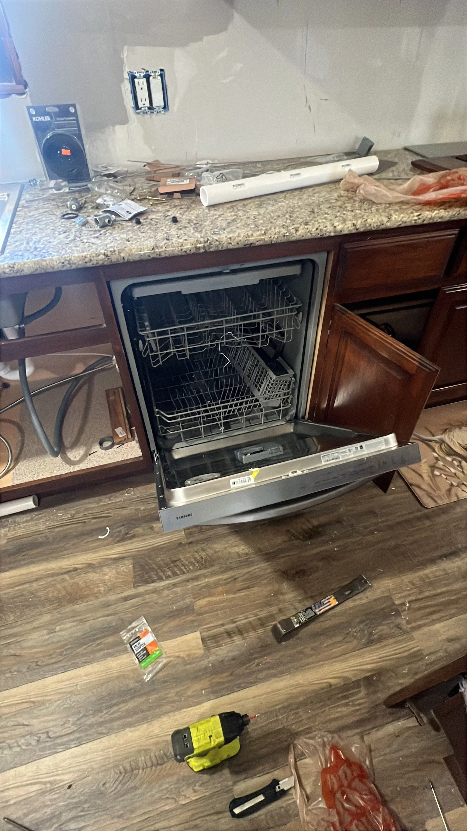 A kitchen countertop with various installation tools, parts, and materials for a dishwasher installation. The dishwasher door is open, and there are loose wires, screws, and packaging on the floor.