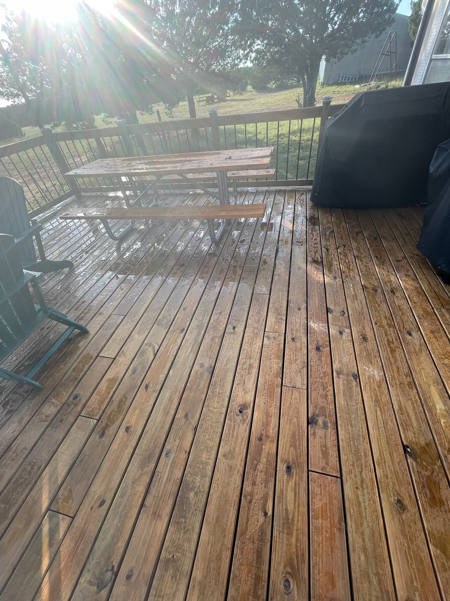 Wooden deck with rain droplets, a black covered grill, a picnic table, and a view of trees and green grassy yard in the background.
