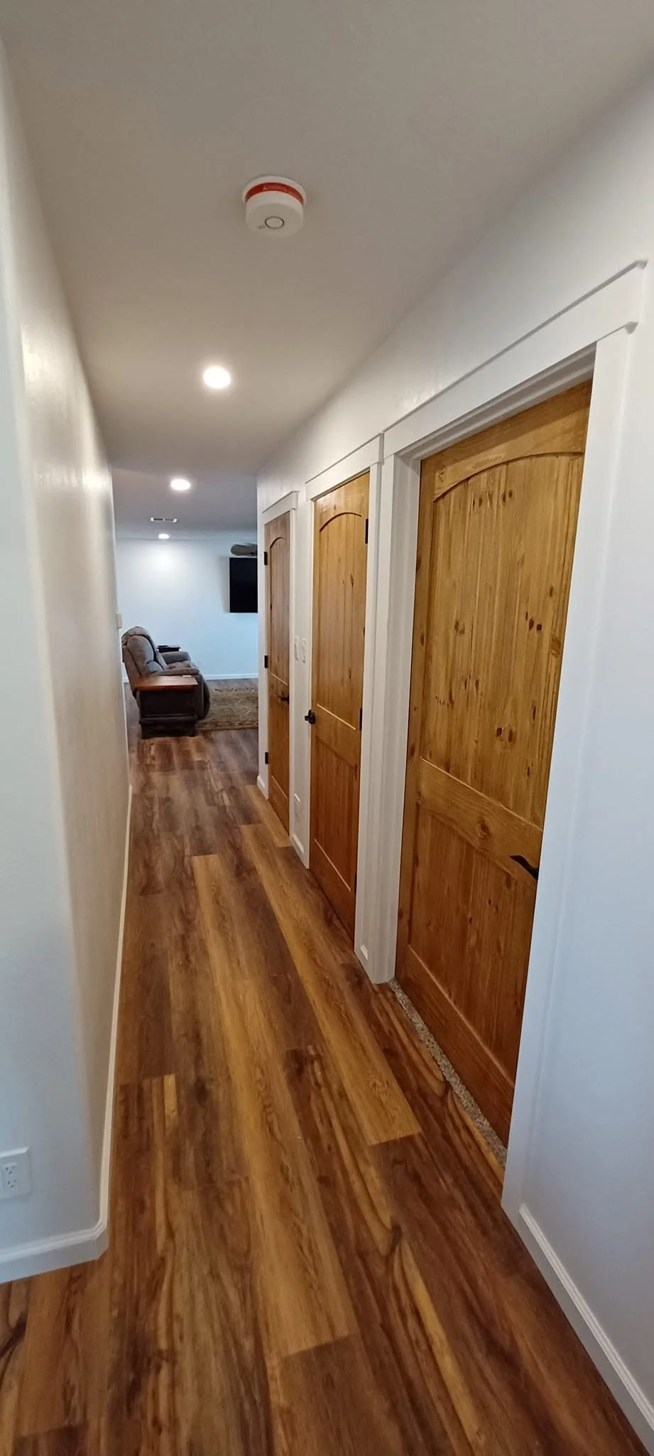 A hallway in a house with three wooden doors on the right side, hardwood floors, white walls, and ceiling with recessed lighting and a smoke detector. At the end of the hall, a living room with two armchairs and a flat-screen TV is visible.