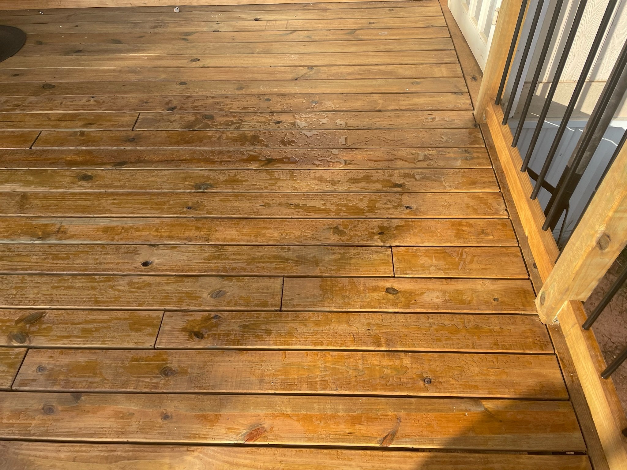 Wooden porch deck with water spots, part of a railing on the right side, and a door in the background.