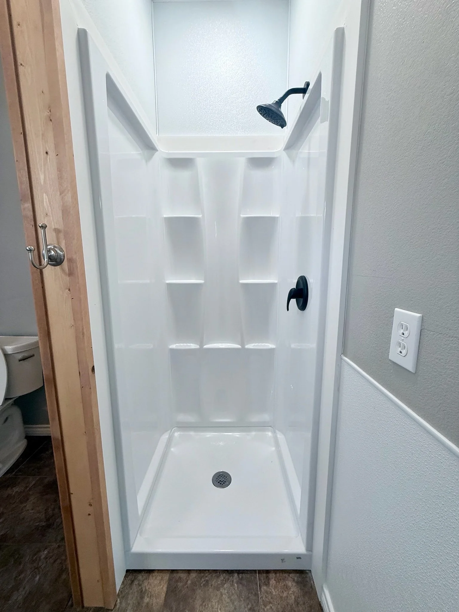 A small, white, enclosed shower with a textured glass door, a black showerhead, built-in shelves on the side, a circular drain at the bottom, and fixtures on the wall.