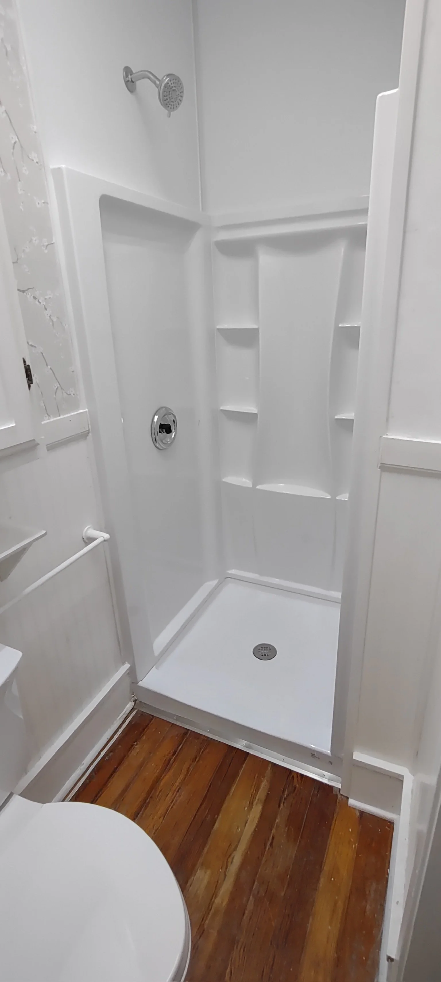 Empty white shower stall with built-in shelves, a showerhead, and a drain, adjacent to a bathroom with wooden floor and nearby toilet.
