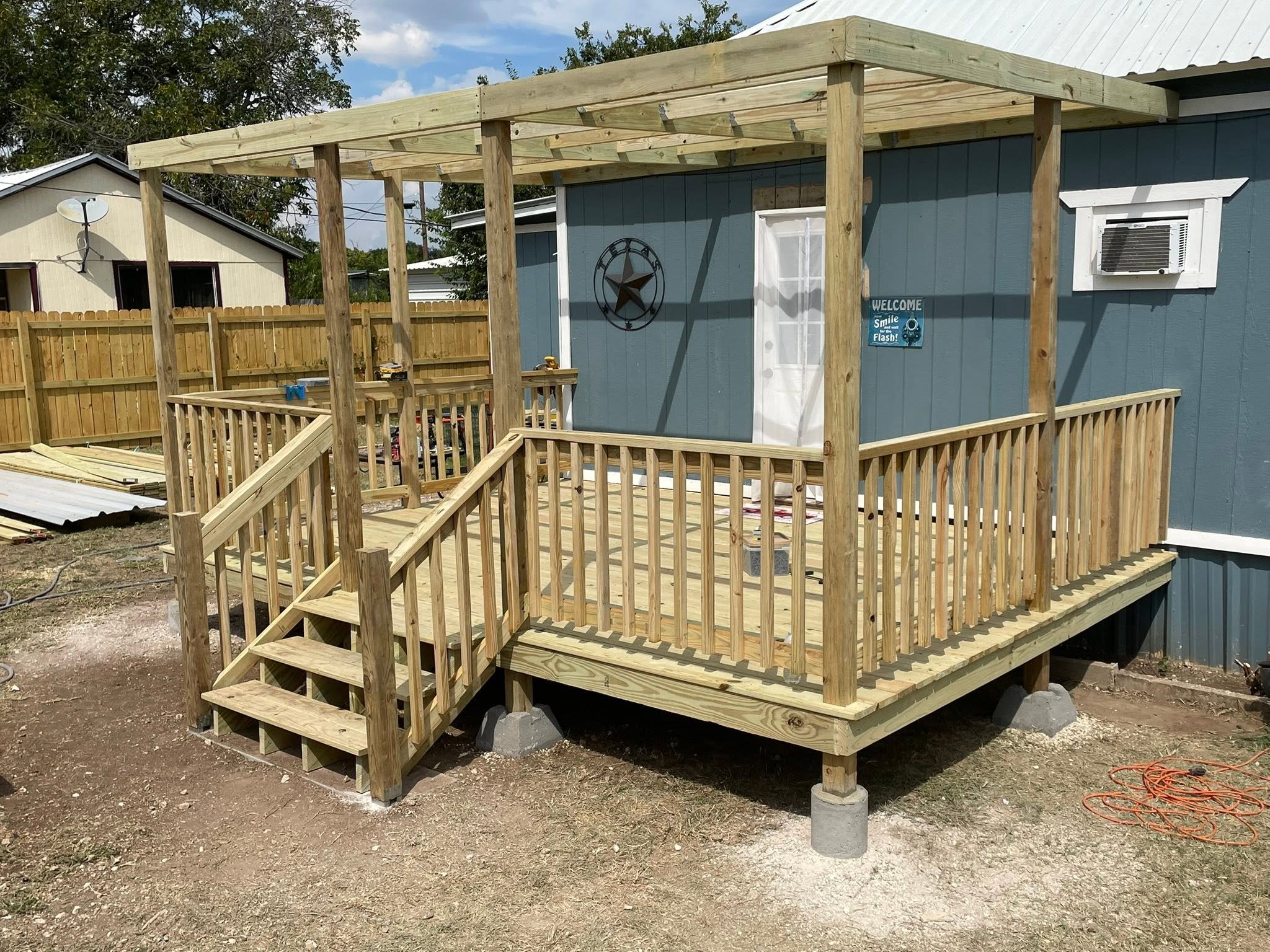 Wooden deck under construction attached to a blue house, with stairs leading down to dirt ground, surrounded by a wooden fence.
