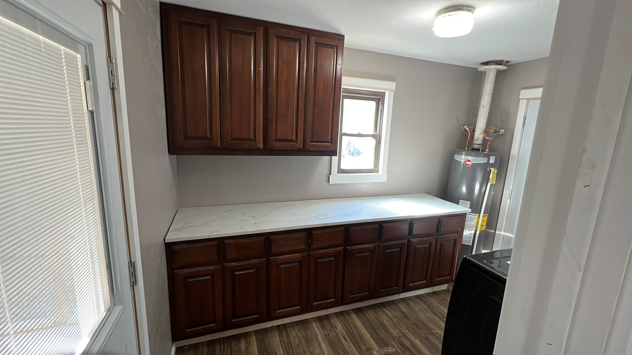 Kitchen with wooden cabinets, a countertop, and a water heater in the corner. There is a window above the countertop and a light fixture on the ceiling.