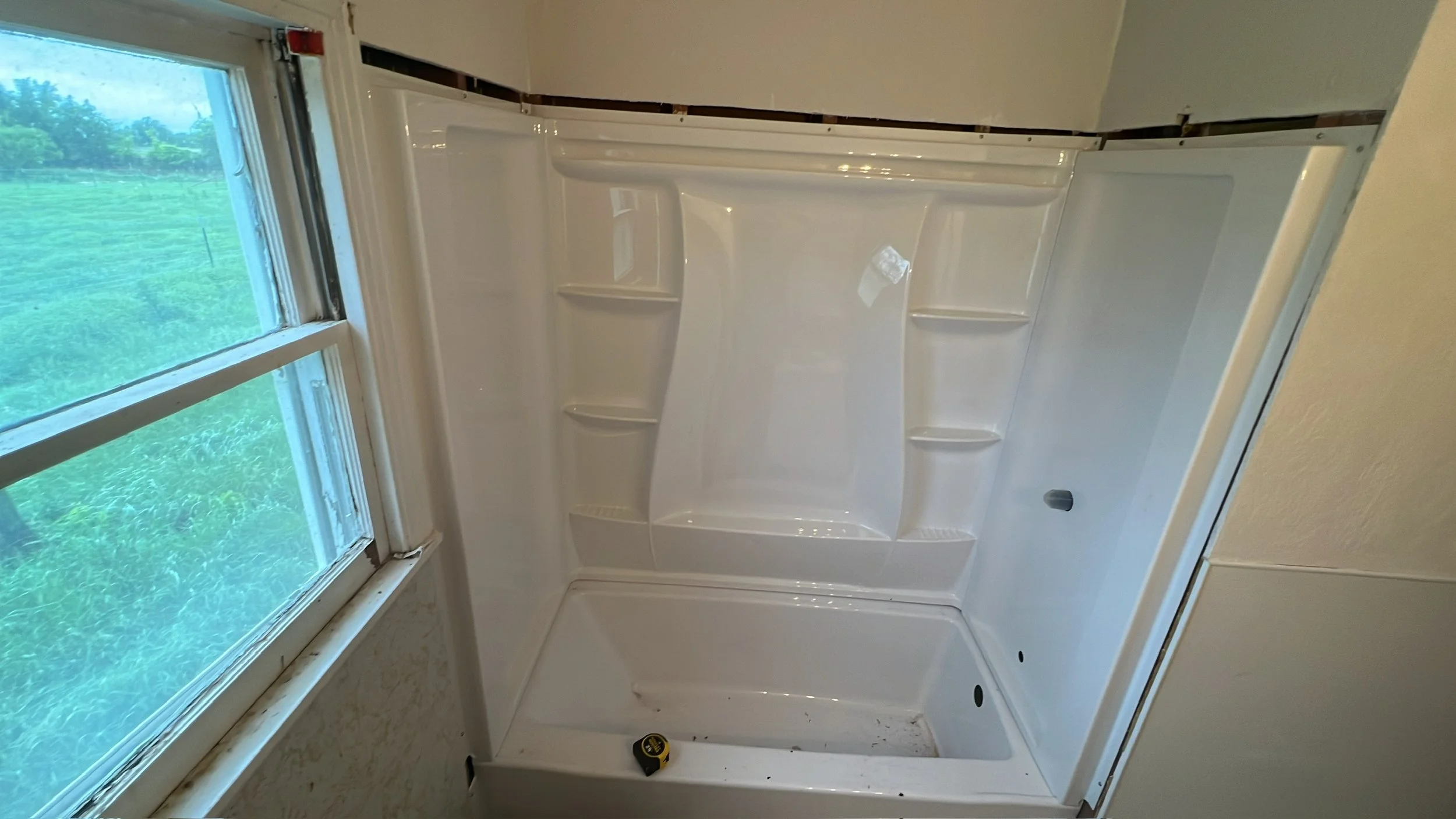 White shower stall with built-in shelves, next to a window showing green foliage outside.