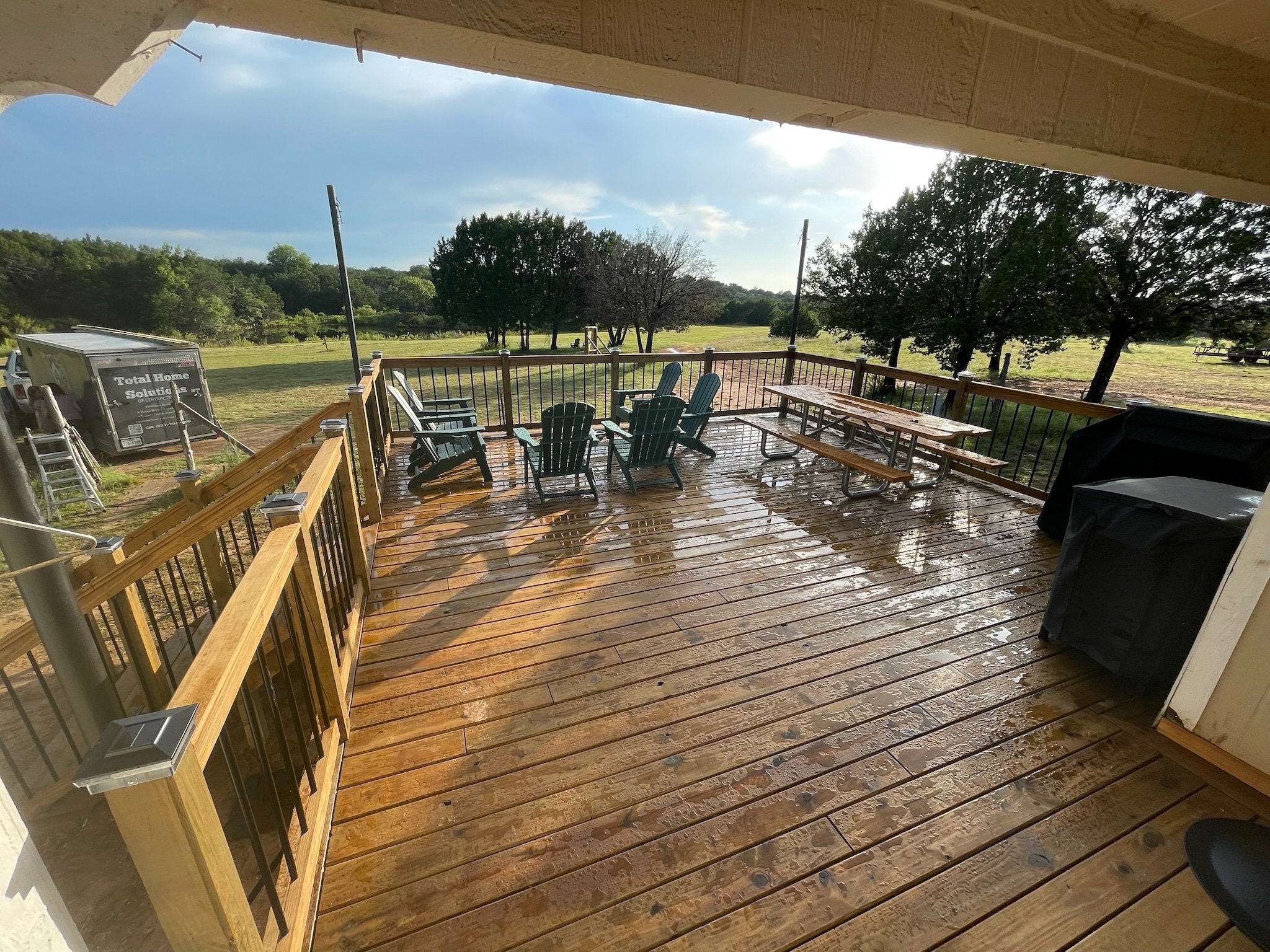 View of a wooden deck with patio furniture, trees, and a grassy open field in the background, viewed from under a covered porch or balcony.