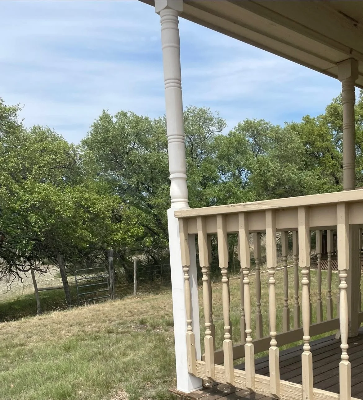 Part of a wooden porch with beige railings and white supporting posts, overlooking a grassy yard with trees and a wire fence under a partly cloudy sky.